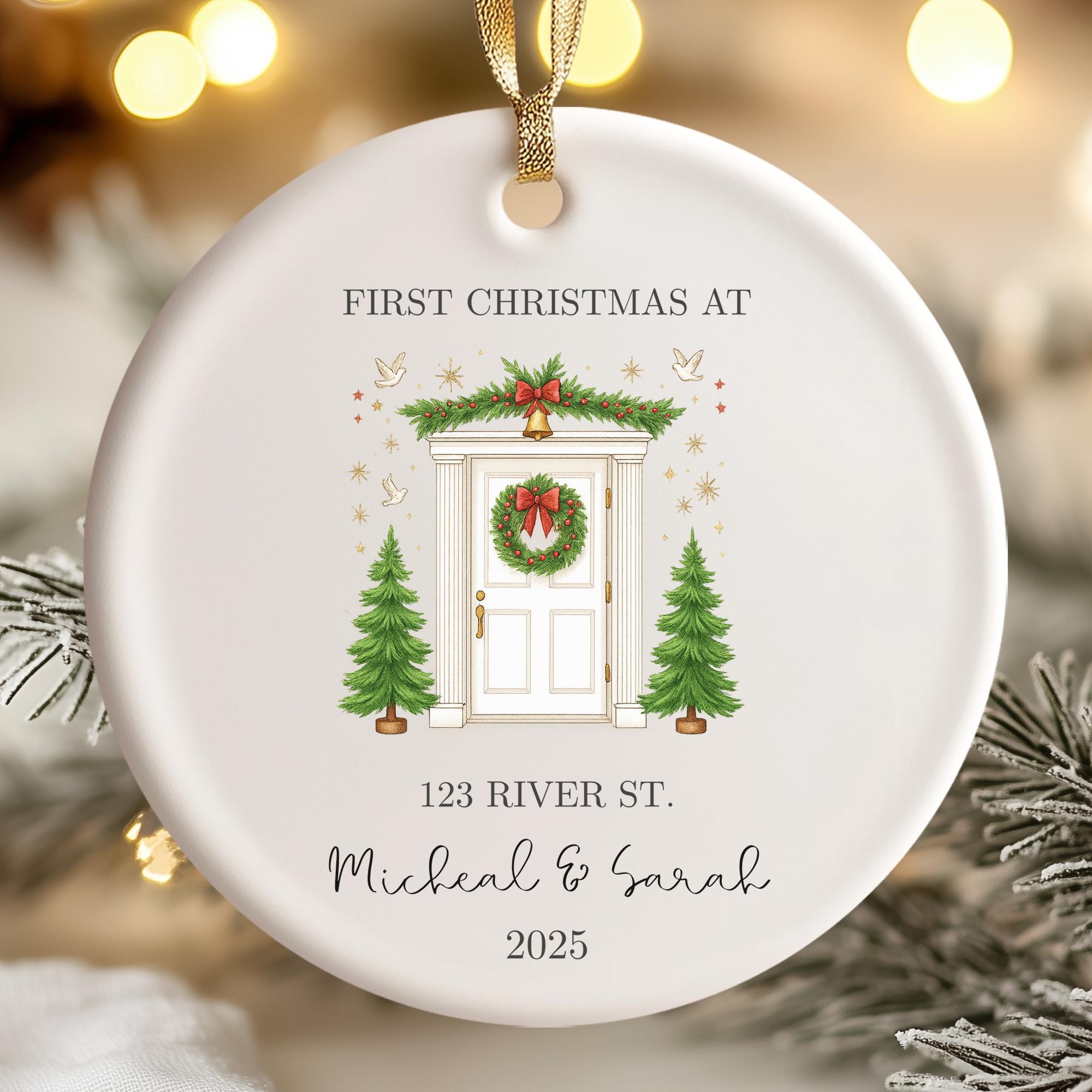 A white Christmas ornament with a wreath, pine trees, and a door, hanging on a string. The ornament has text that reads "FIRST CHRISTMAS AT 123 RIVER ST. Michael & Sarah" and "2025".