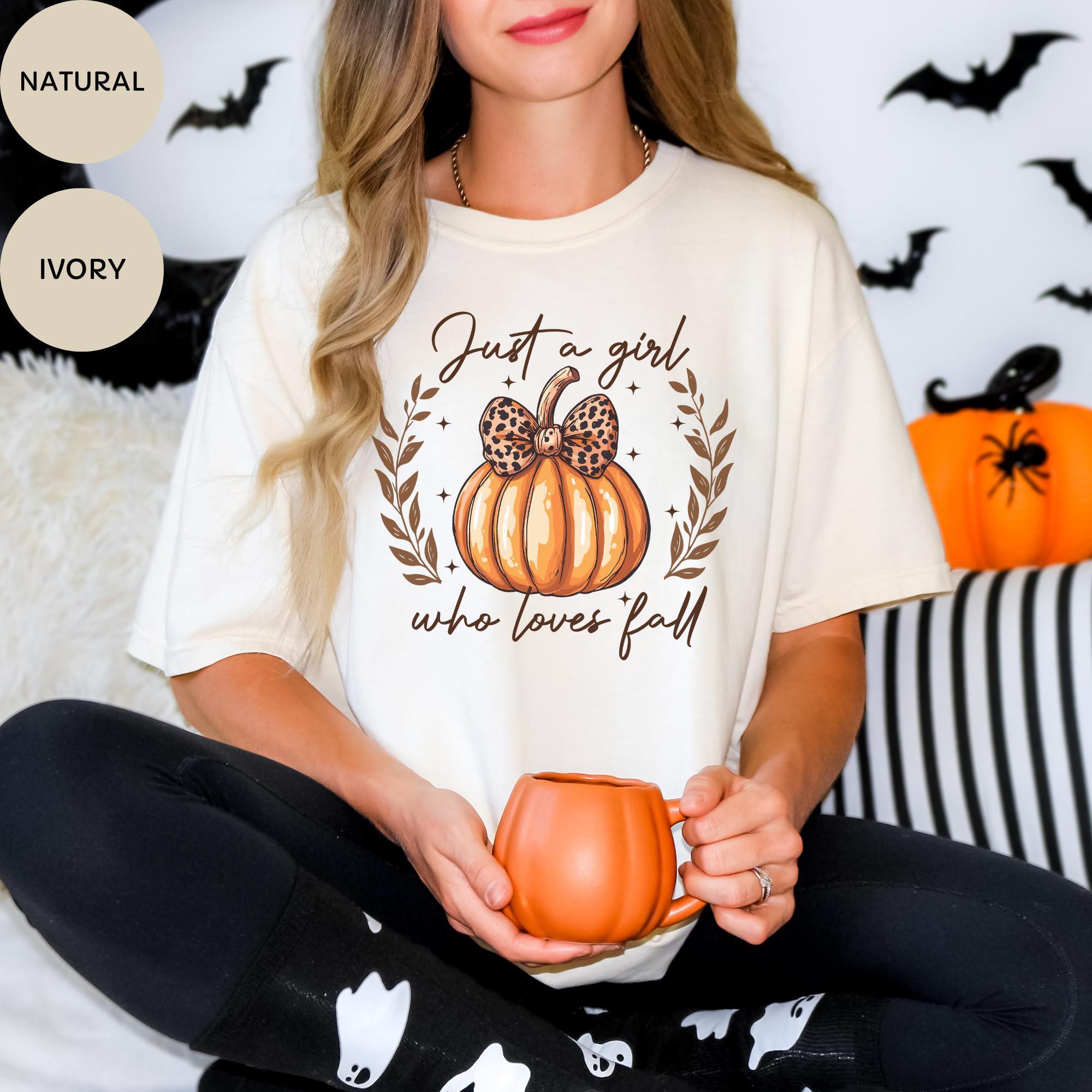 A woman wearing a white t-shirt with a pumpkin graphic and the text "Just a girl who loves fall" is sitting on a couch with a mug in her hand.