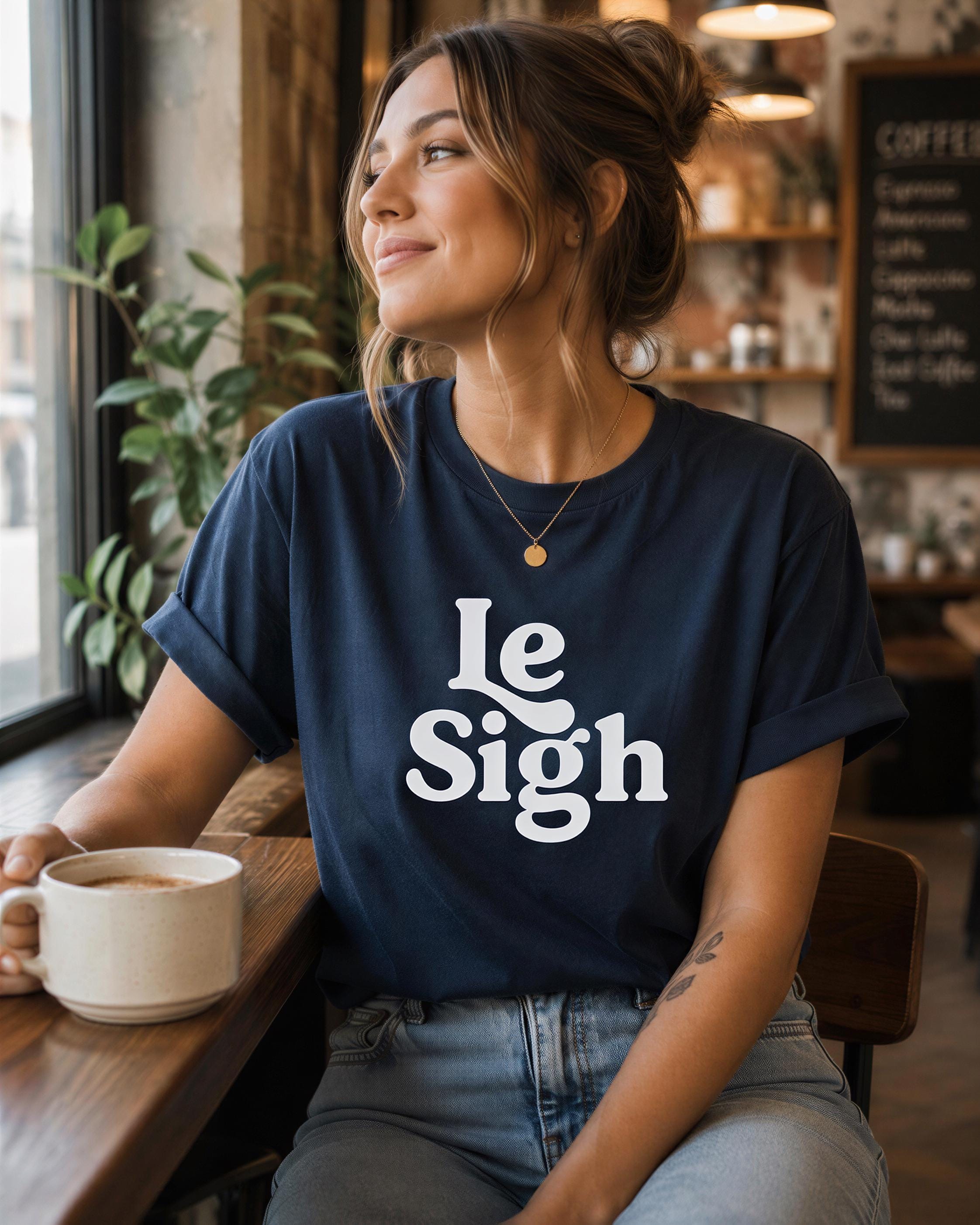 A woman wearing a navy blue t-shirt with the text "Le Sigh" is sitting at a table in a cafe, holding a coffee mug and looking out the window.