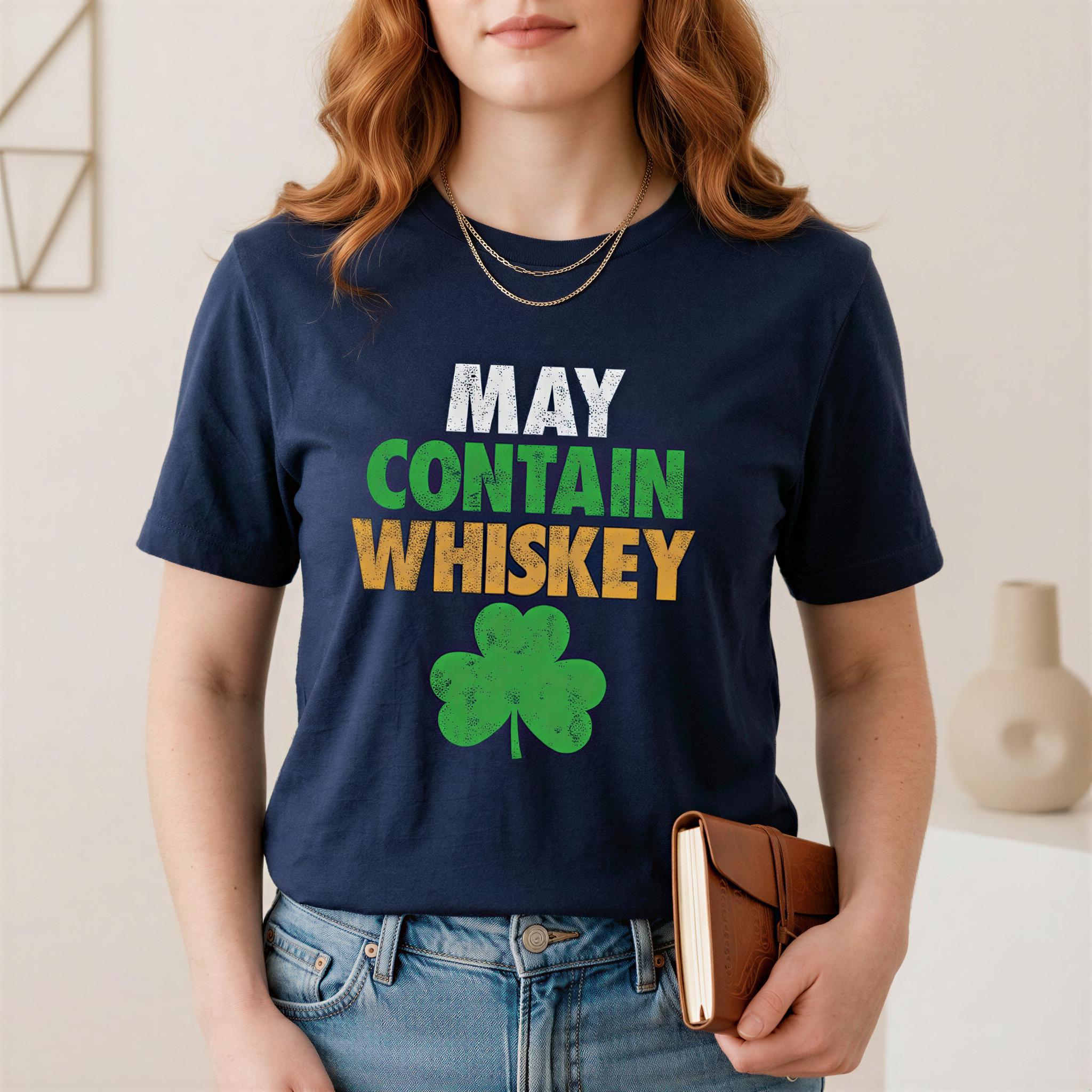 A woman wearing a navy blue t-shirt with the text "MAY CONTAIN WHISKEY" printed on it, along with a green shamrock graphic. She is holding a brown leather-bound notebook in her hand.
