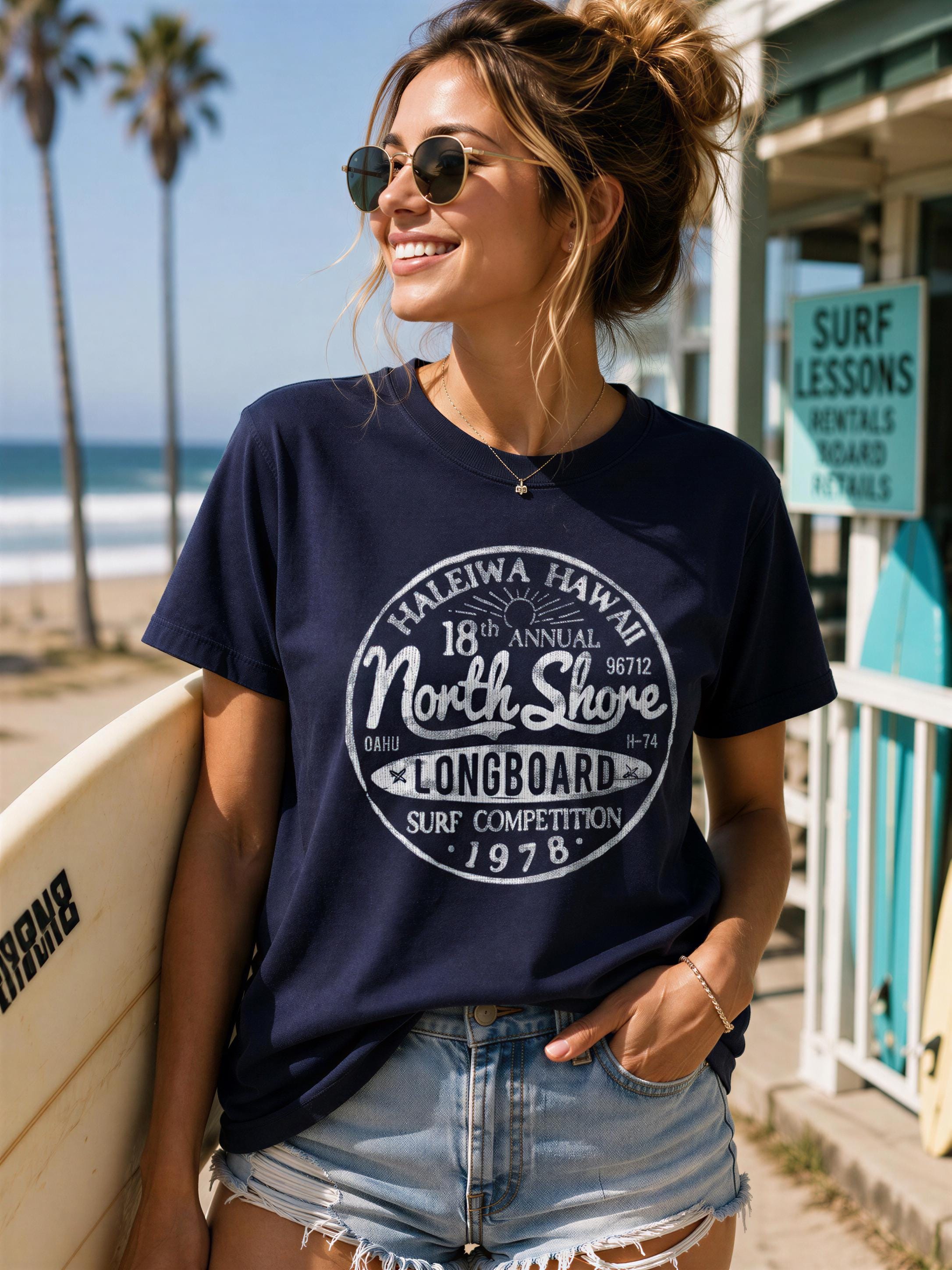 A woman wearing a navy blue t-shirt with a graphic design, sunglasses, and denim shorts is holding a surfboard and smiling at the camera.