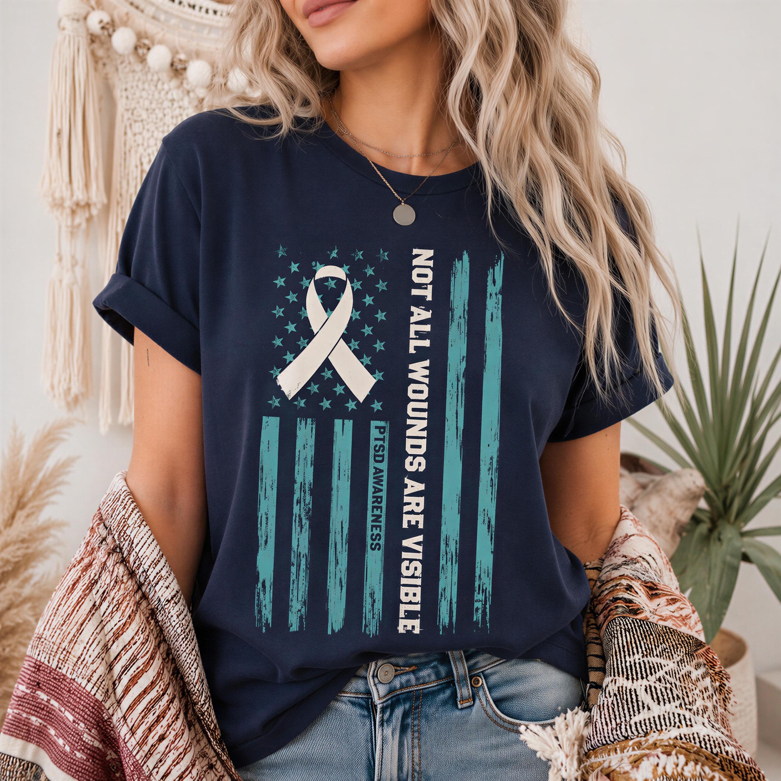 A woman wearing a navy blue t-shirt with a graphic design featuring a white ribbon and an American flag, along with the text "NOT ALL WOUNDS ARE VISIBLE" printed on it.