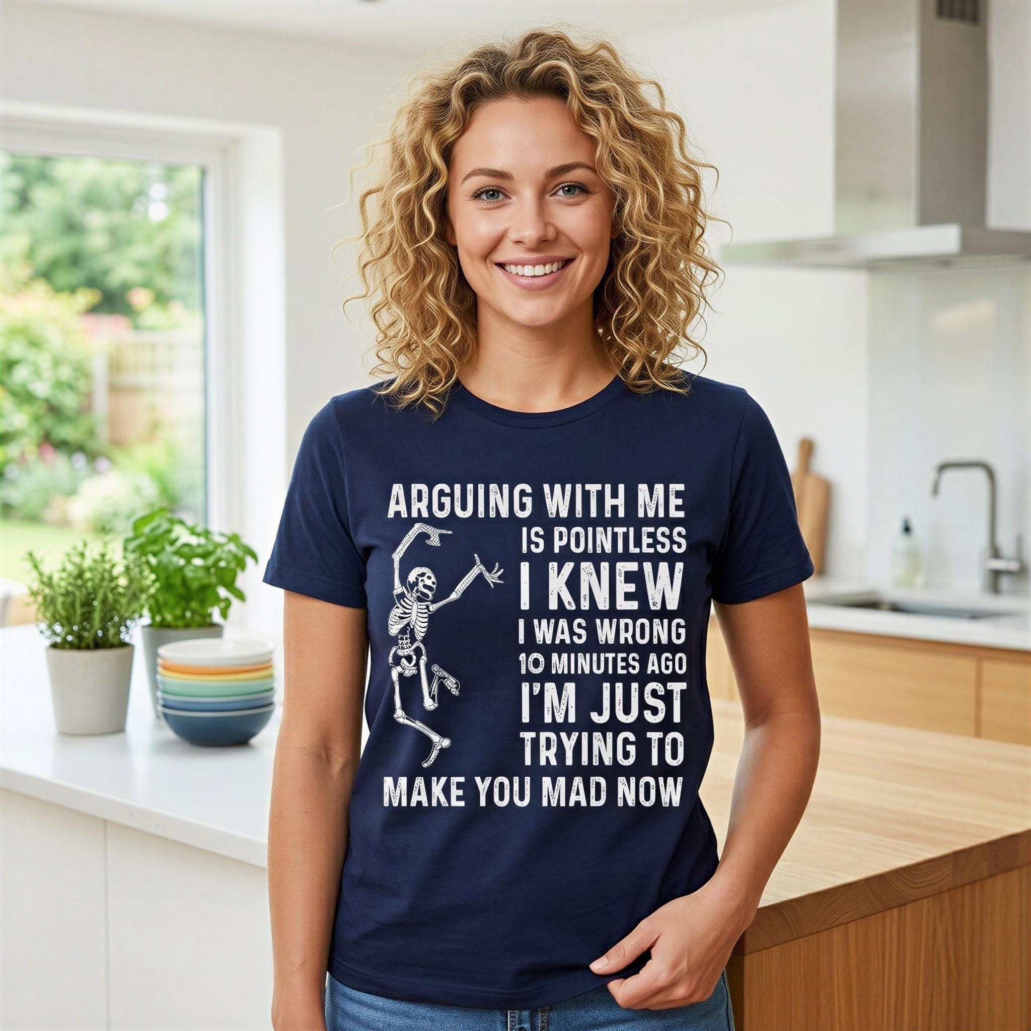 A woman with curly blonde hair wearing a navy blue t-shirt with a humorous text printed on it, standing in a kitchen.