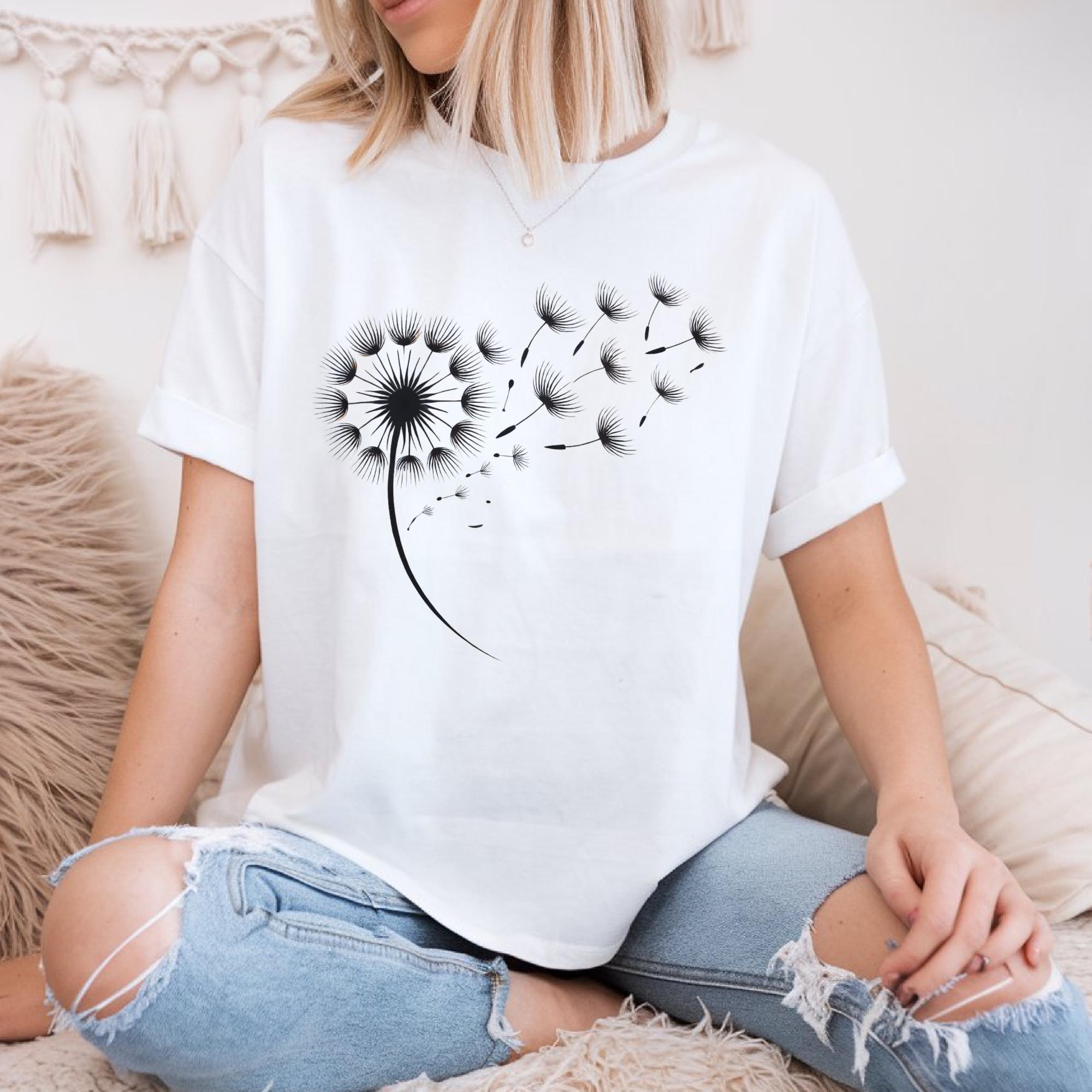 A woman wearing a white t-shirt with a black graphic design of a dandelion blowing in the wind, sitting on a couch.