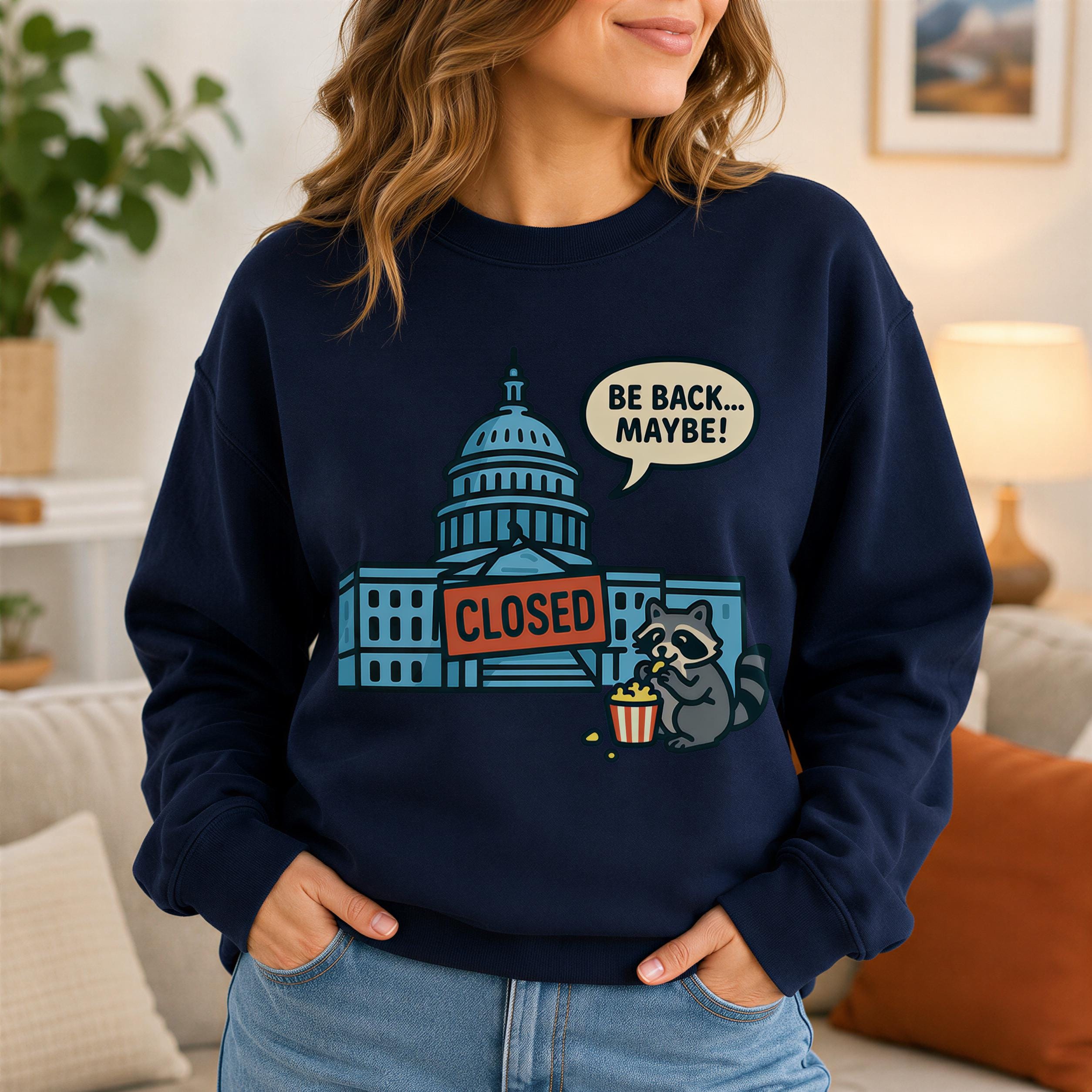 A woman wearing a navy blue sweatshirt with a graphic design depicting a raccoon holding a sign that says "CLOSED" and a speech bubble saying "BE BACK MAYBE!"