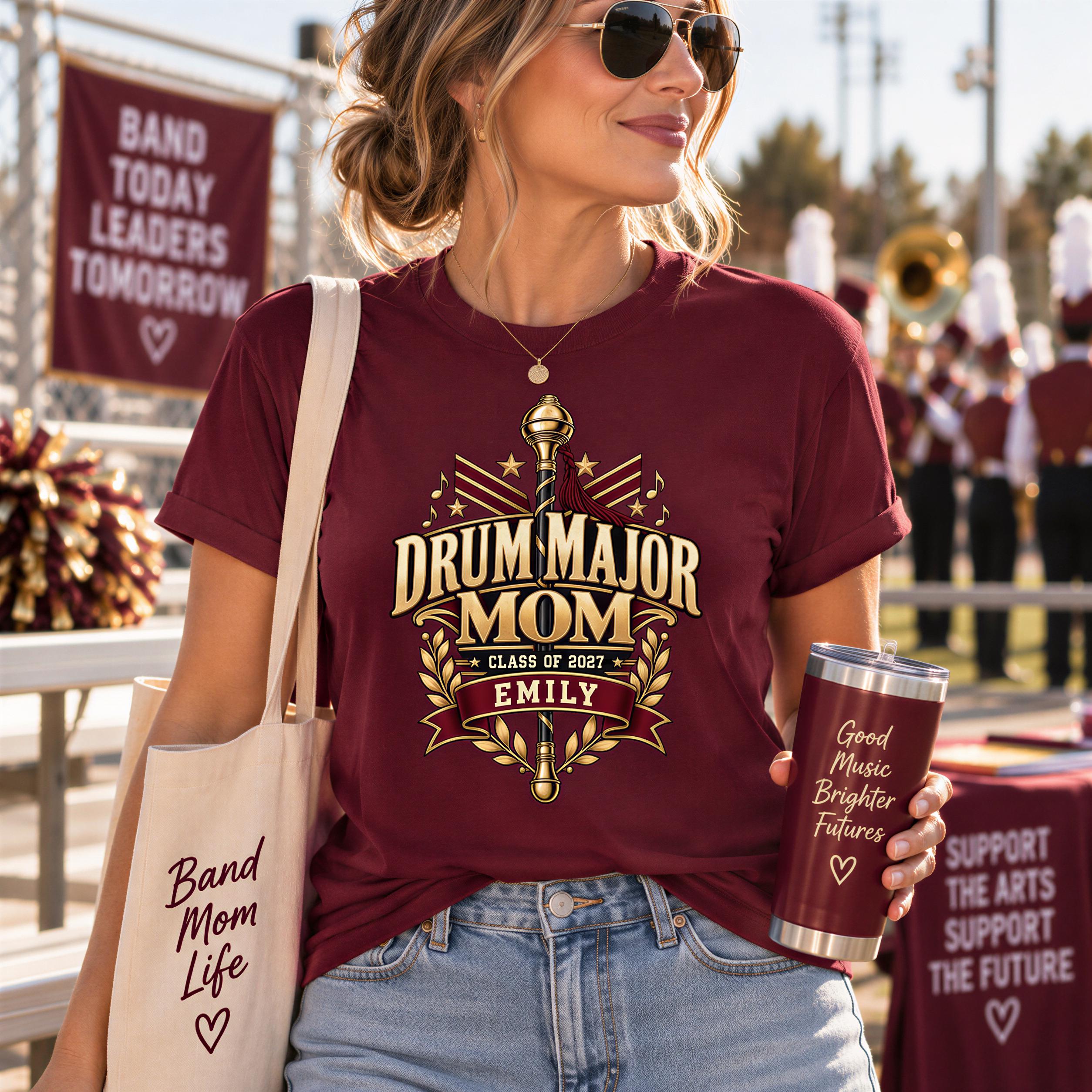 A woman wearing a burgundy t-shirt with a drum major logo and holding a red tumbler with text, standing in front of a marching band.