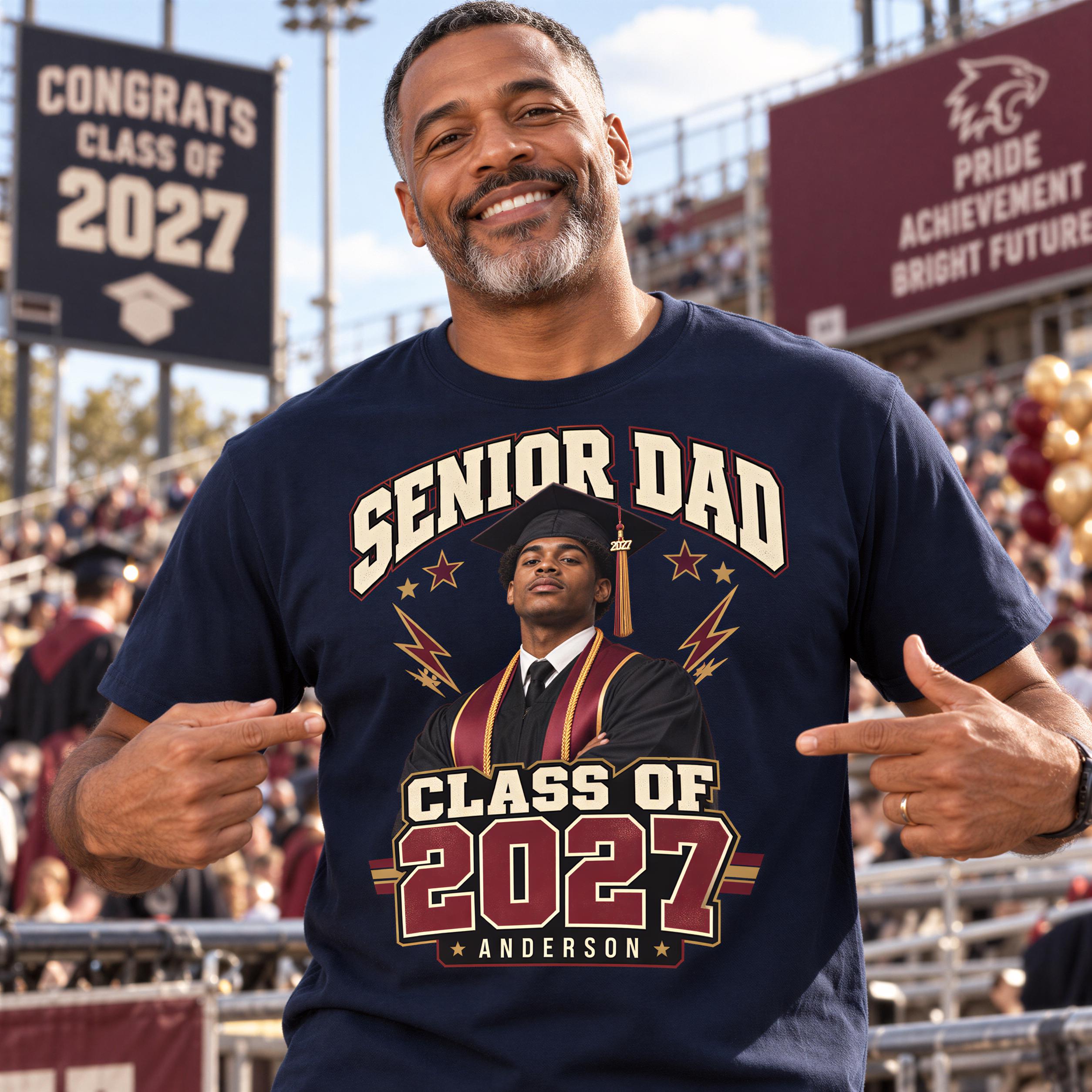 A smiling man wearing a t-shirt with a graphic of a graduation cap and the text "SENIOR DAD CLASS OF 2027 ANDERSON" pointing at the shirt.