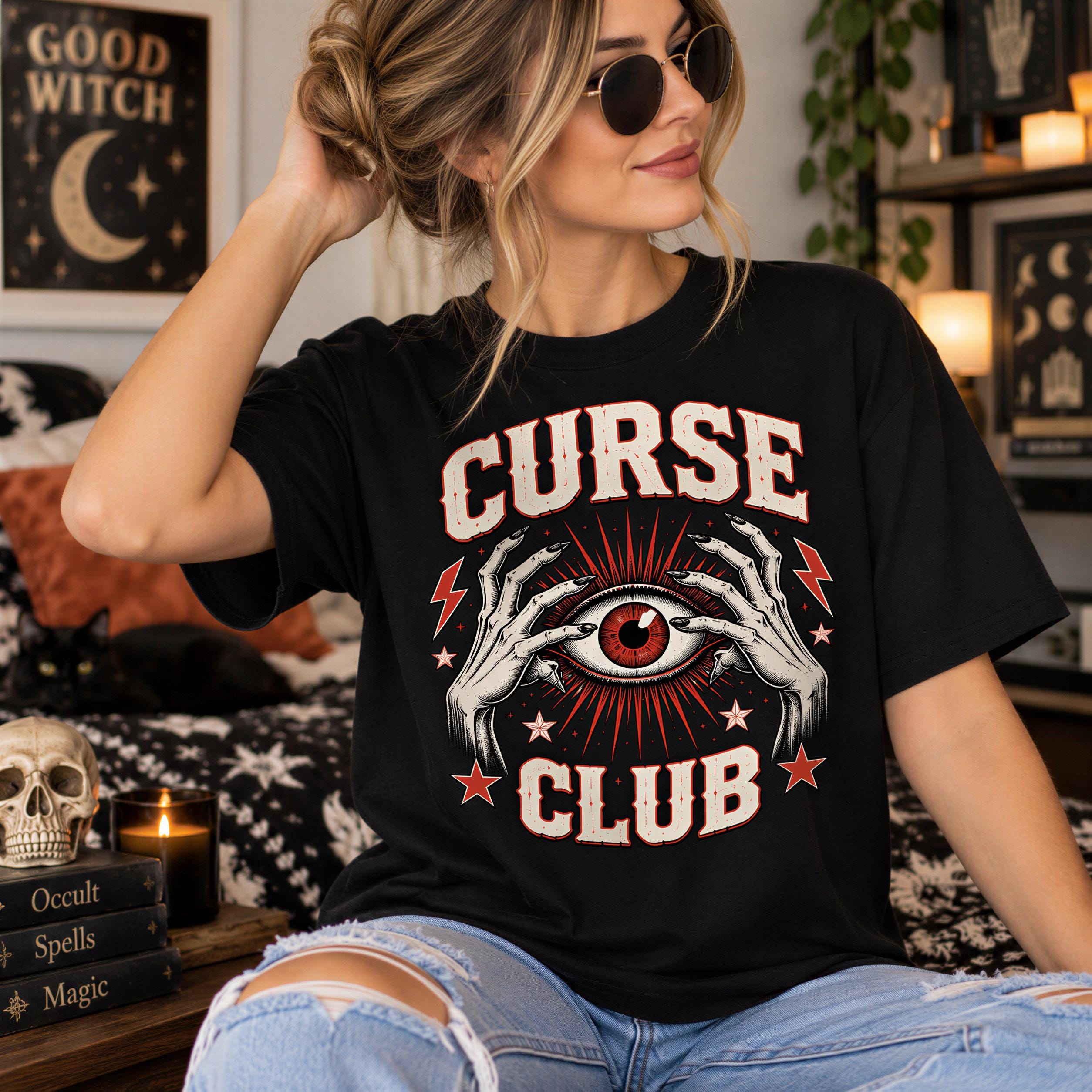 A woman wearing a black t-shirt with a graphic design of a red eye and hands, sitting on a bed with a skull and candles in the background.