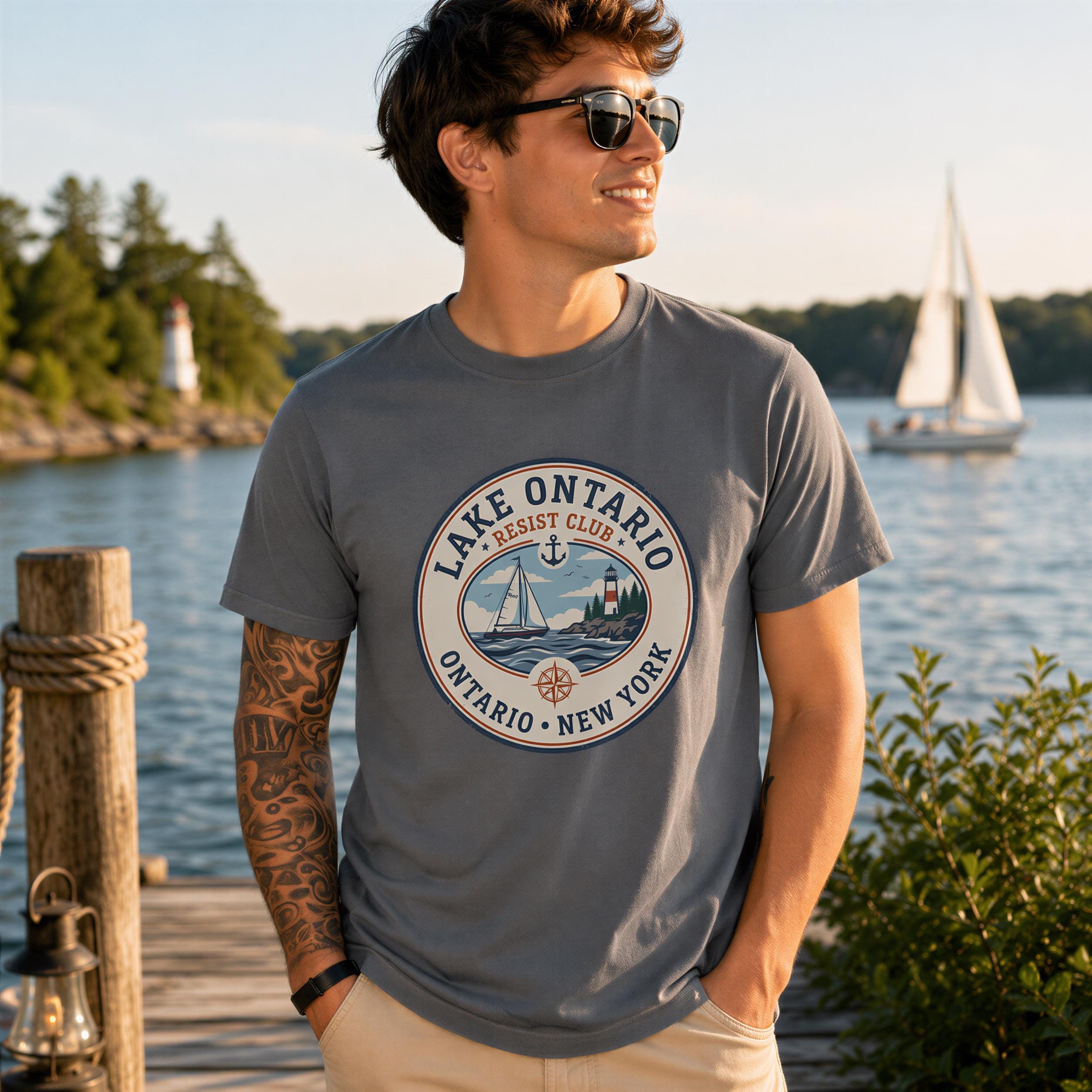 A man wearing a grey t-shirt with a graphic design stands near a wooden dock, with a sailboat visible in the background.