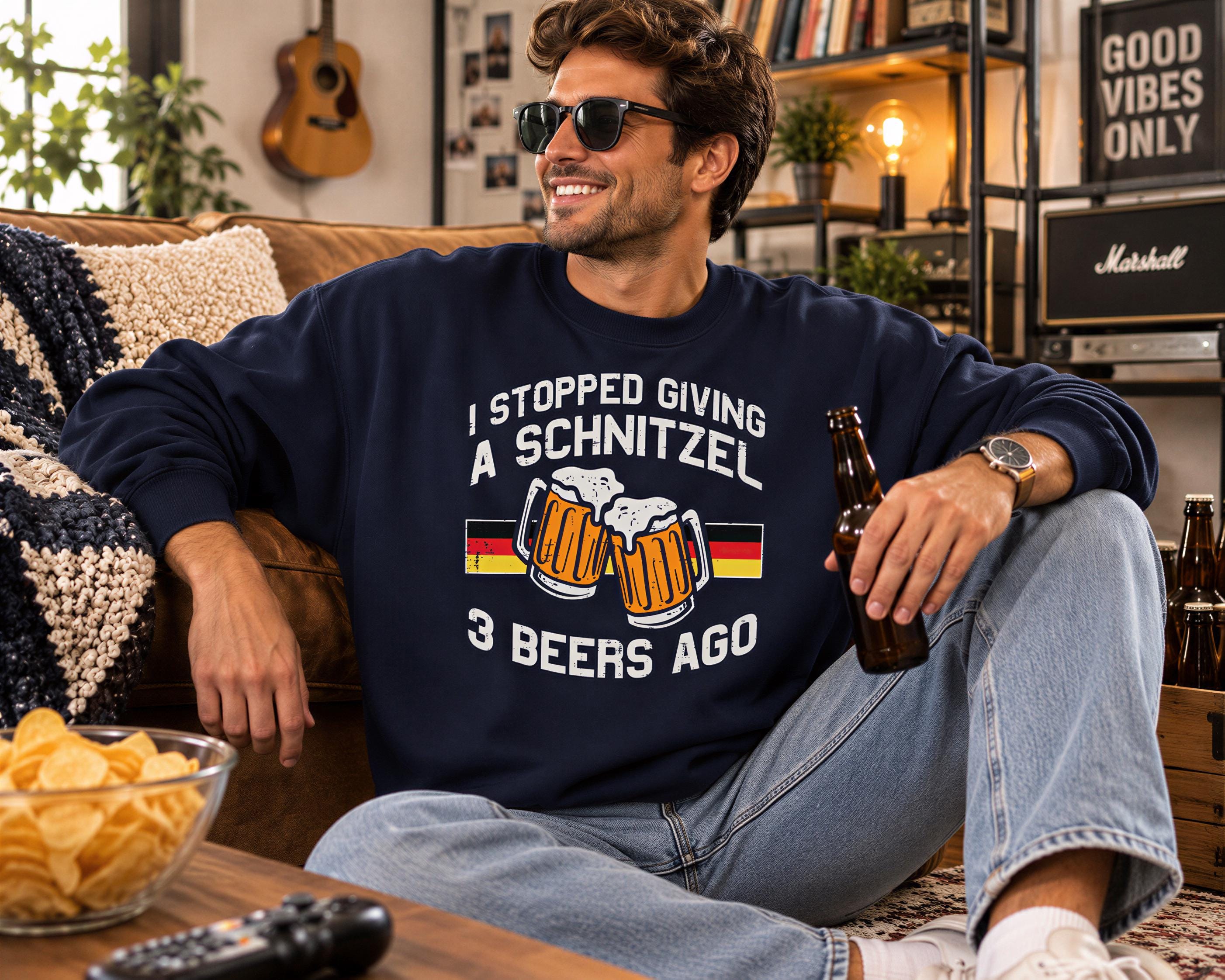 A man wearing a blue sweatshirt with a humorous text and graphic, sitting on a couch and holding a beer bottle.