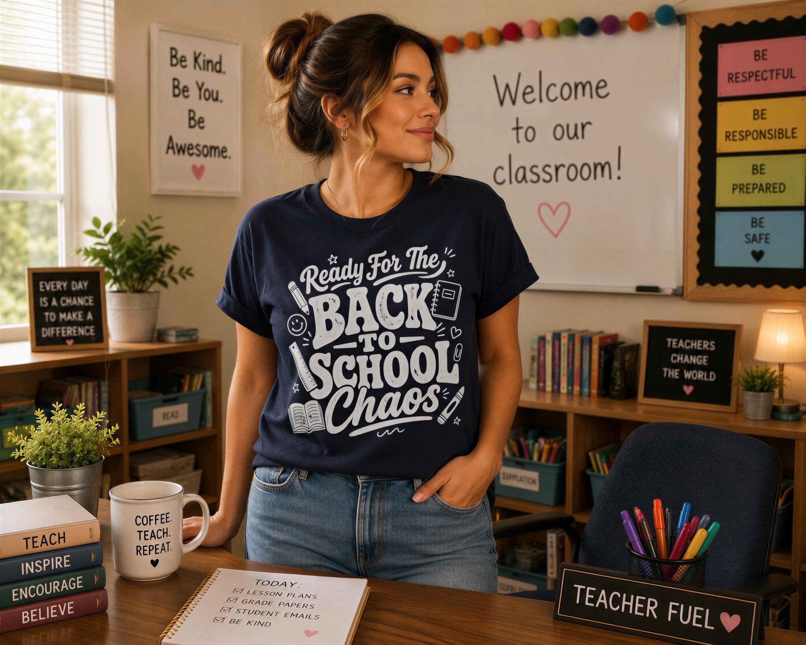A woman stands in a classroom, wearing a blue t-shirt with the text "Back to School Chaos" and smiling. The classroom has a whiteboard, bookshelves, and a sign that says "Welcome to our classroom!"