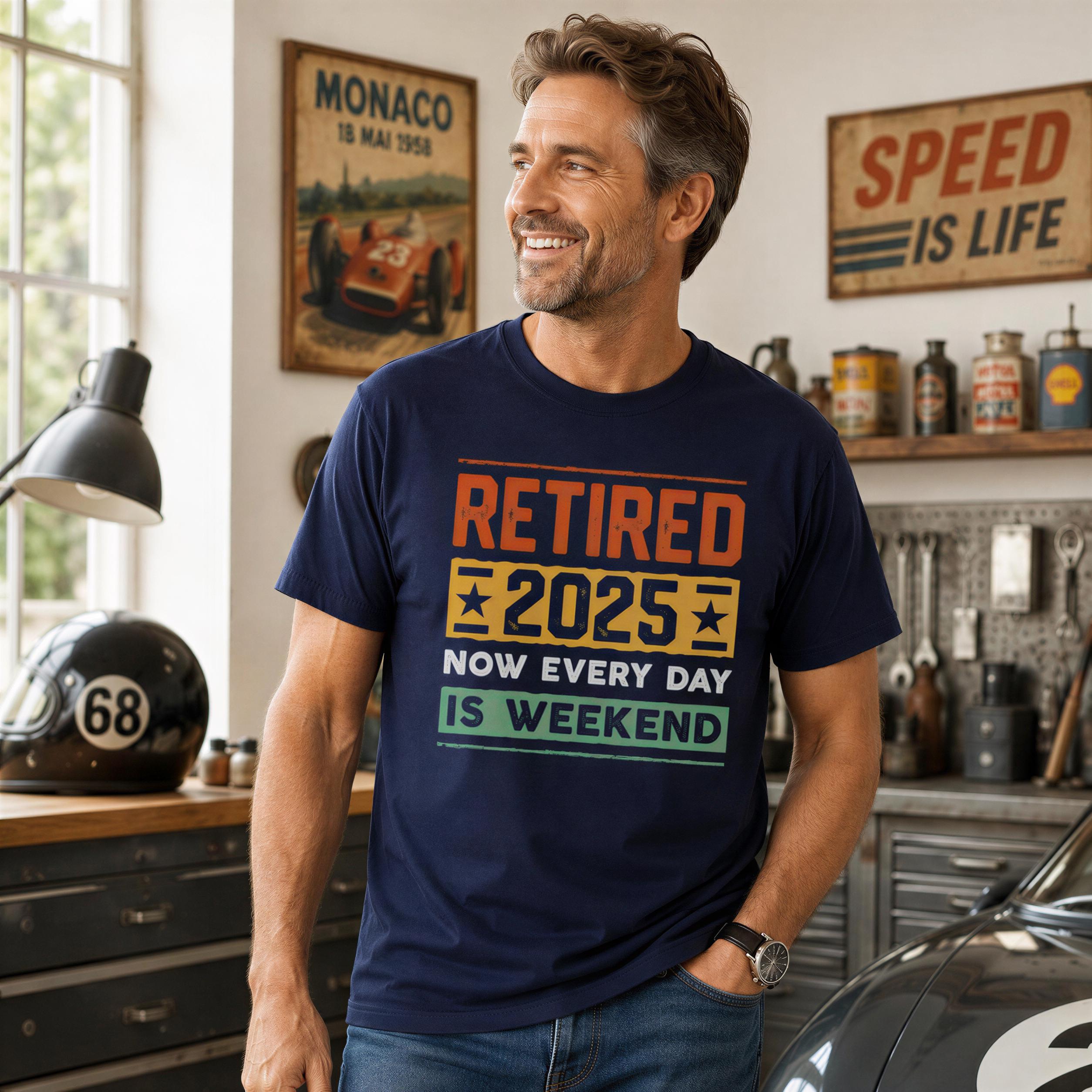 A man wearing a blue t-shirt with text and a graphic of a race car is smiling and standing in a garage with various tools and equipment visible in the background.