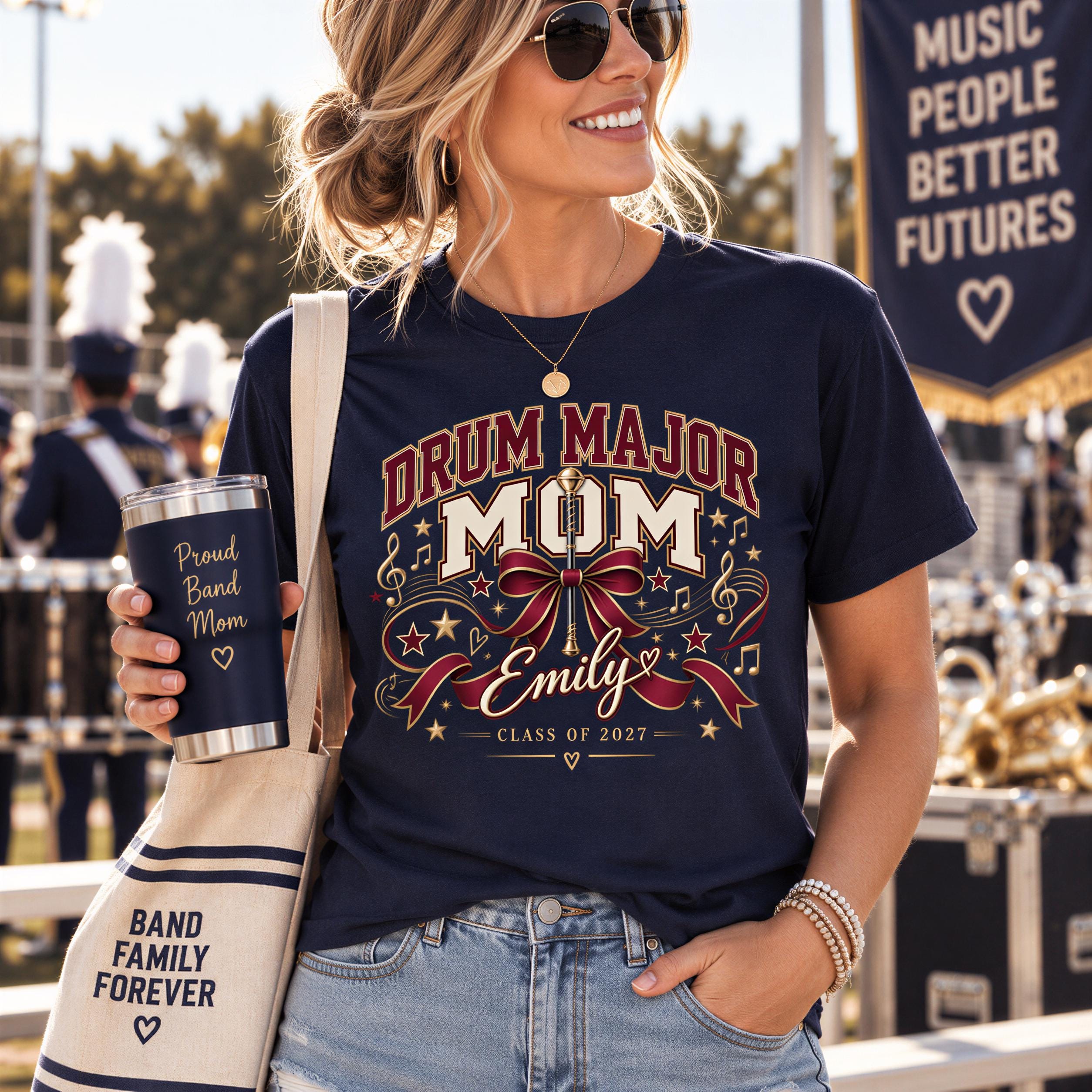 A woman wearing a navy blue t-shirt with a drum major design and holding a black tumbler with a message that says "Pride Band Mom Emily Class of 2027" is standing in front of a marching band.