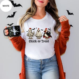May include: White t-shirt with a Halloween graphic of three cartoon chickens dressed as a mummy, ghost, and vampire. The text "Chick or Treat" is below the chickens. The model is wearing blue jeans and an orange cardigan.