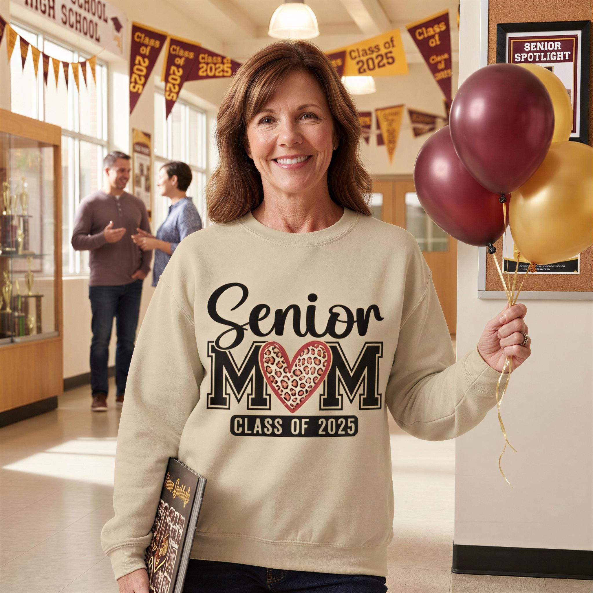 A smiling woman wearing a sweatshirt with the text "SENIOR MOM CLASS OF 2025" holding balloons in a hallway with other people present.