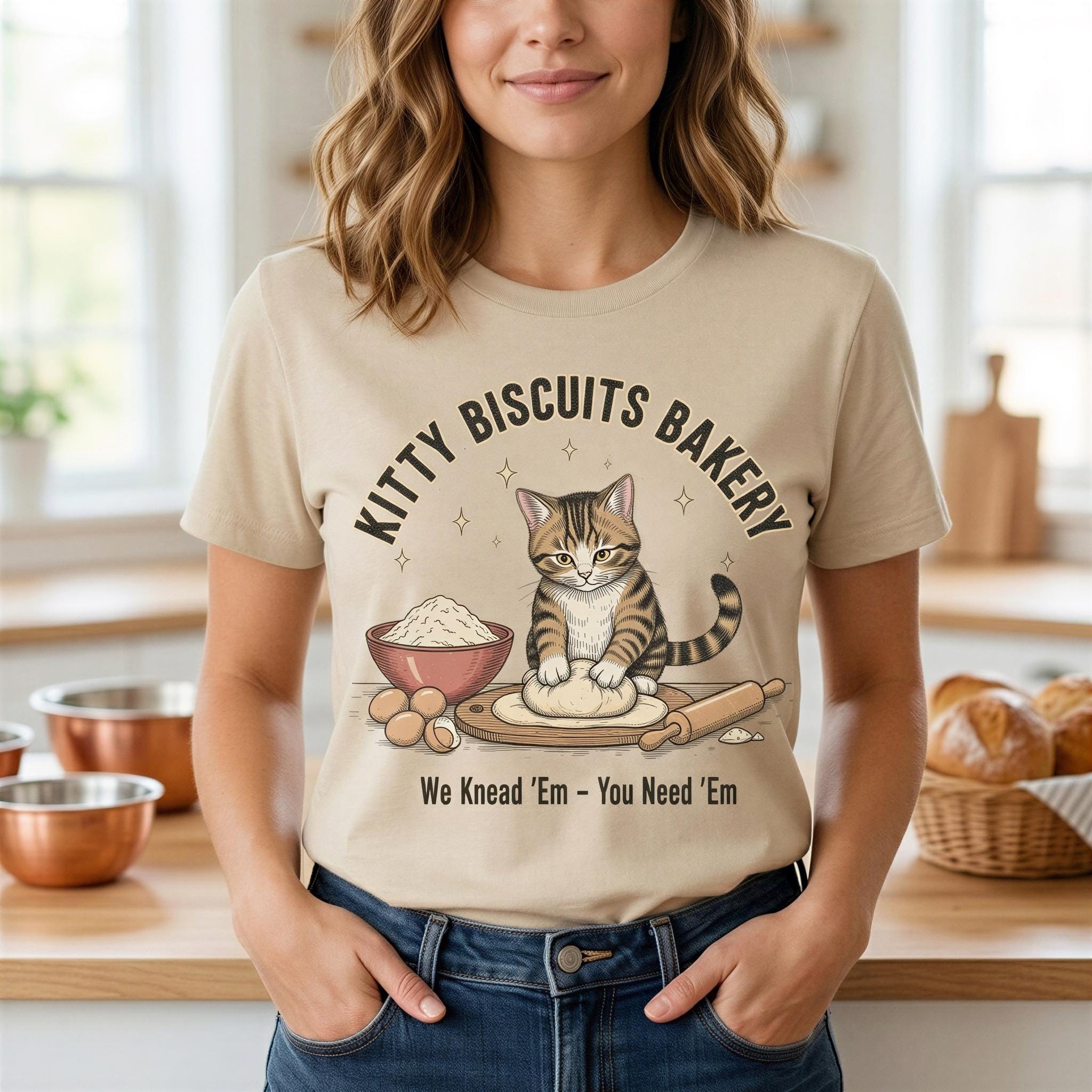 A woman wearing a t-shirt with a graphic design of a cat making biscuits, surrounded by kitchen items such as bowls and a rolling pin.