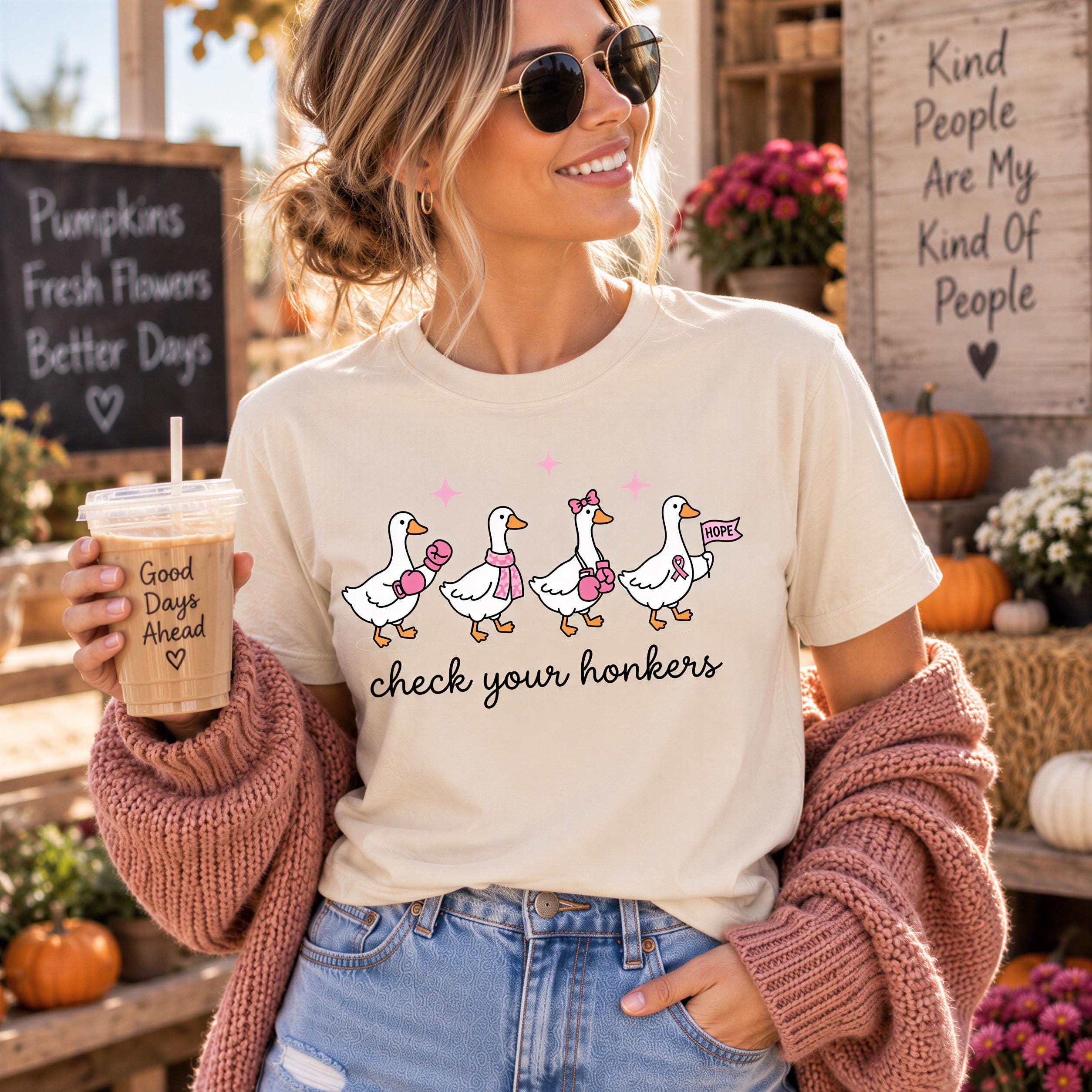 A woman wearing a white t-shirt with a cartoon of ducks and holding a drink stands in front of a chalkboard sign and a wooden sign with a message about kindness.