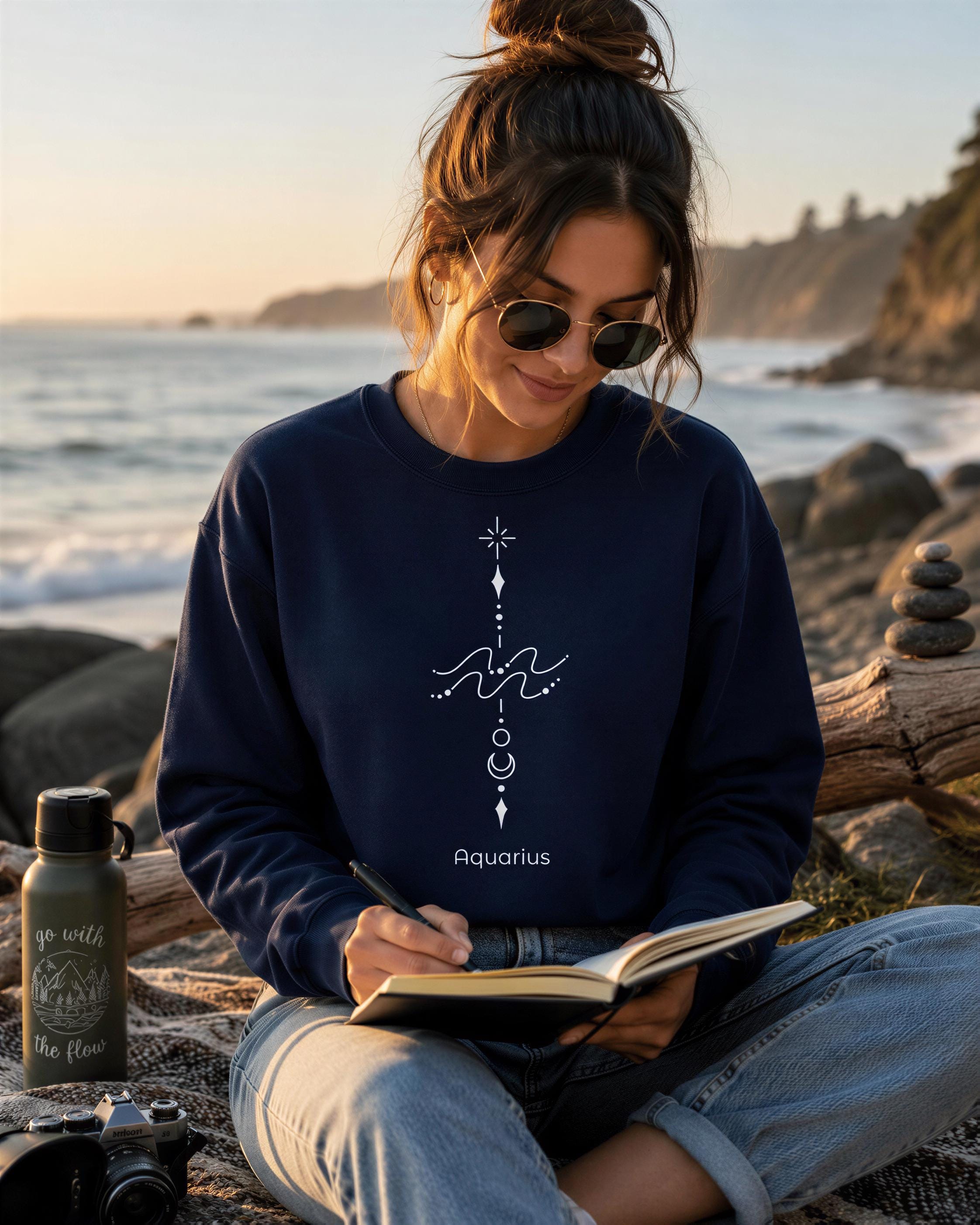 A woman wearing a navy blue sweatshirt with a white design sits on a blanket by the ocean, writing in a notebook.