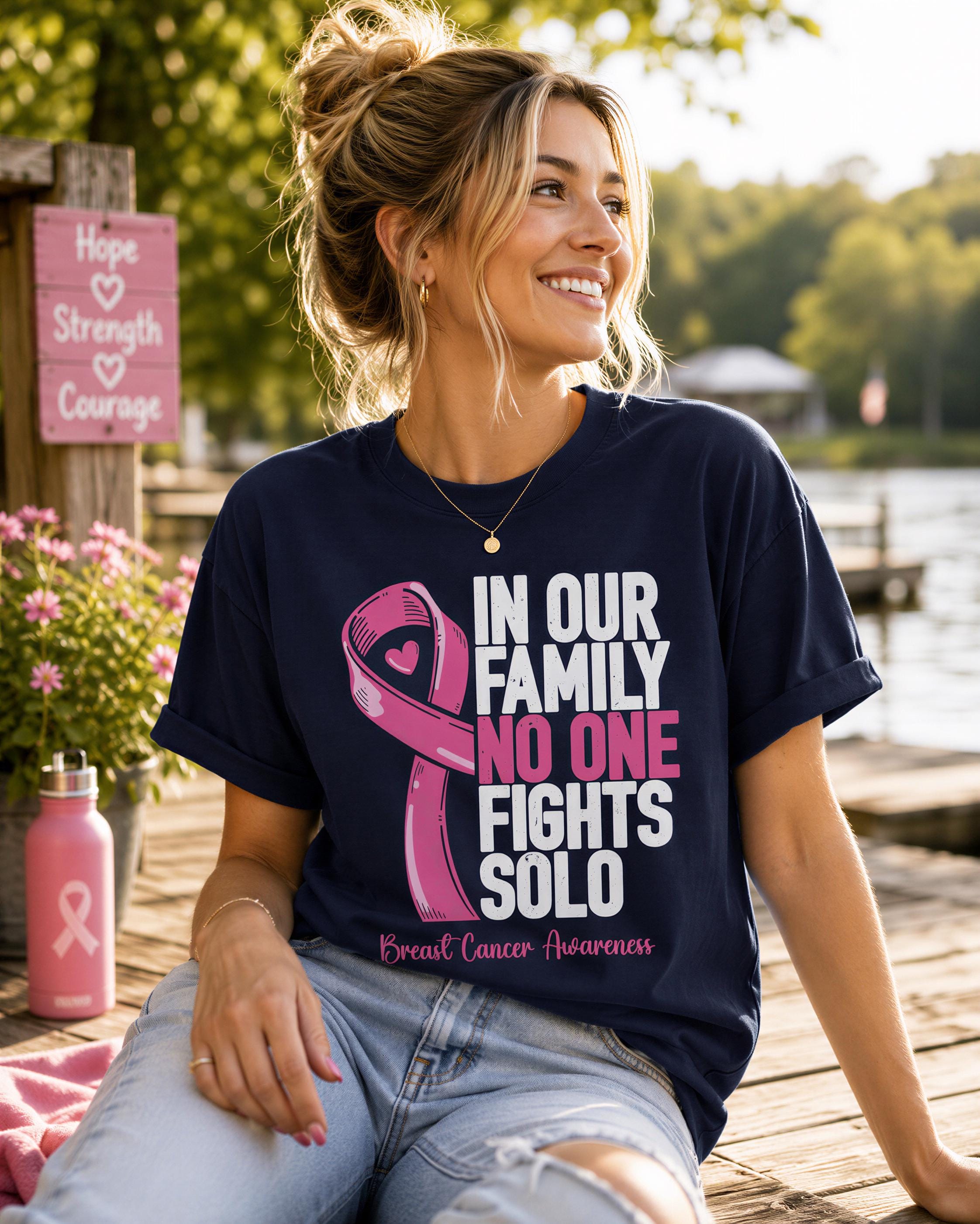 A woman wearing a black t-shirt with pink text and a pink ribbon design, sitting on a wooden bench near a body of water.