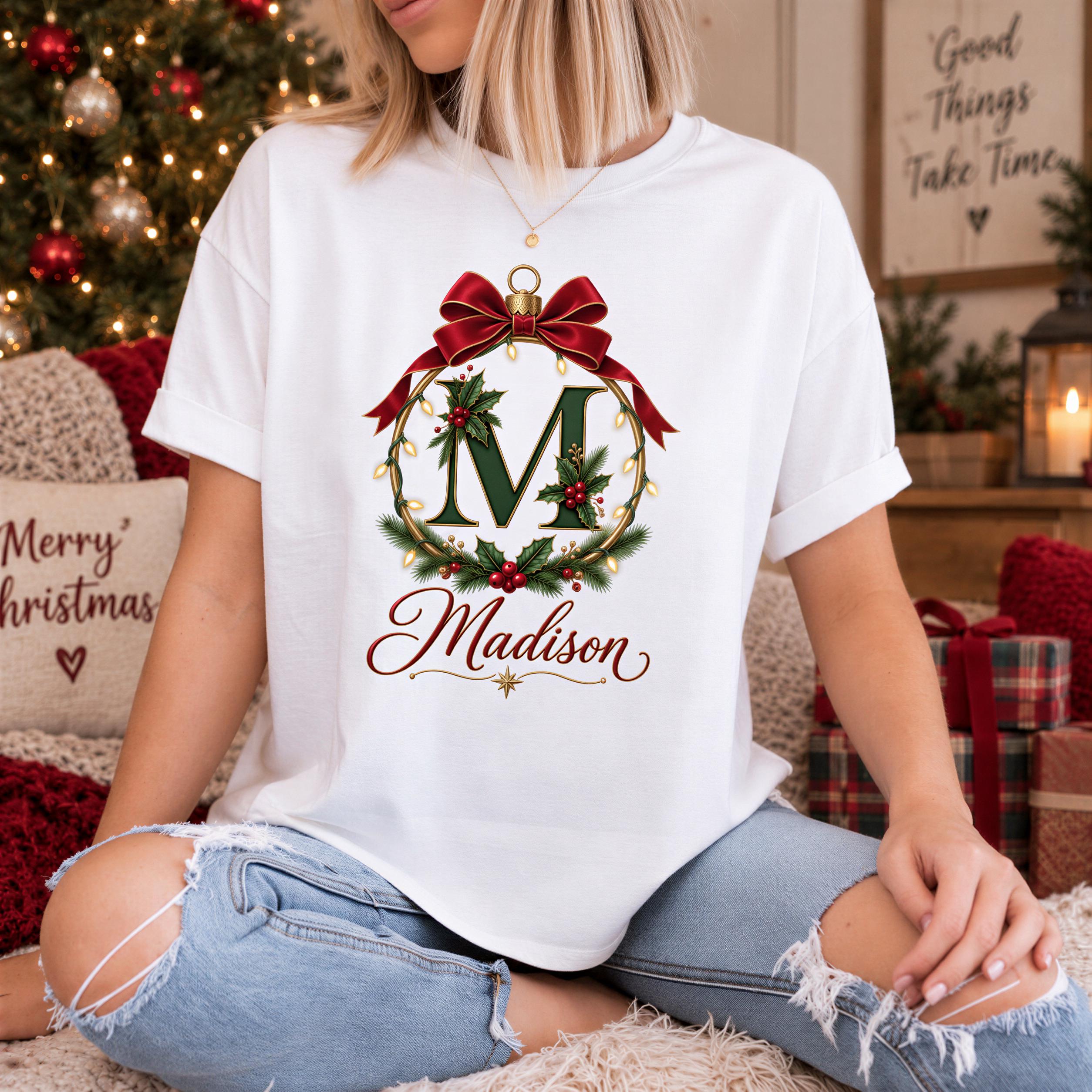 A woman wearing a white t-shirt with a graphic design of a Christmas ornament and the name "Madison" printed on it, sitting on a bed with Christmas decorations in the background.