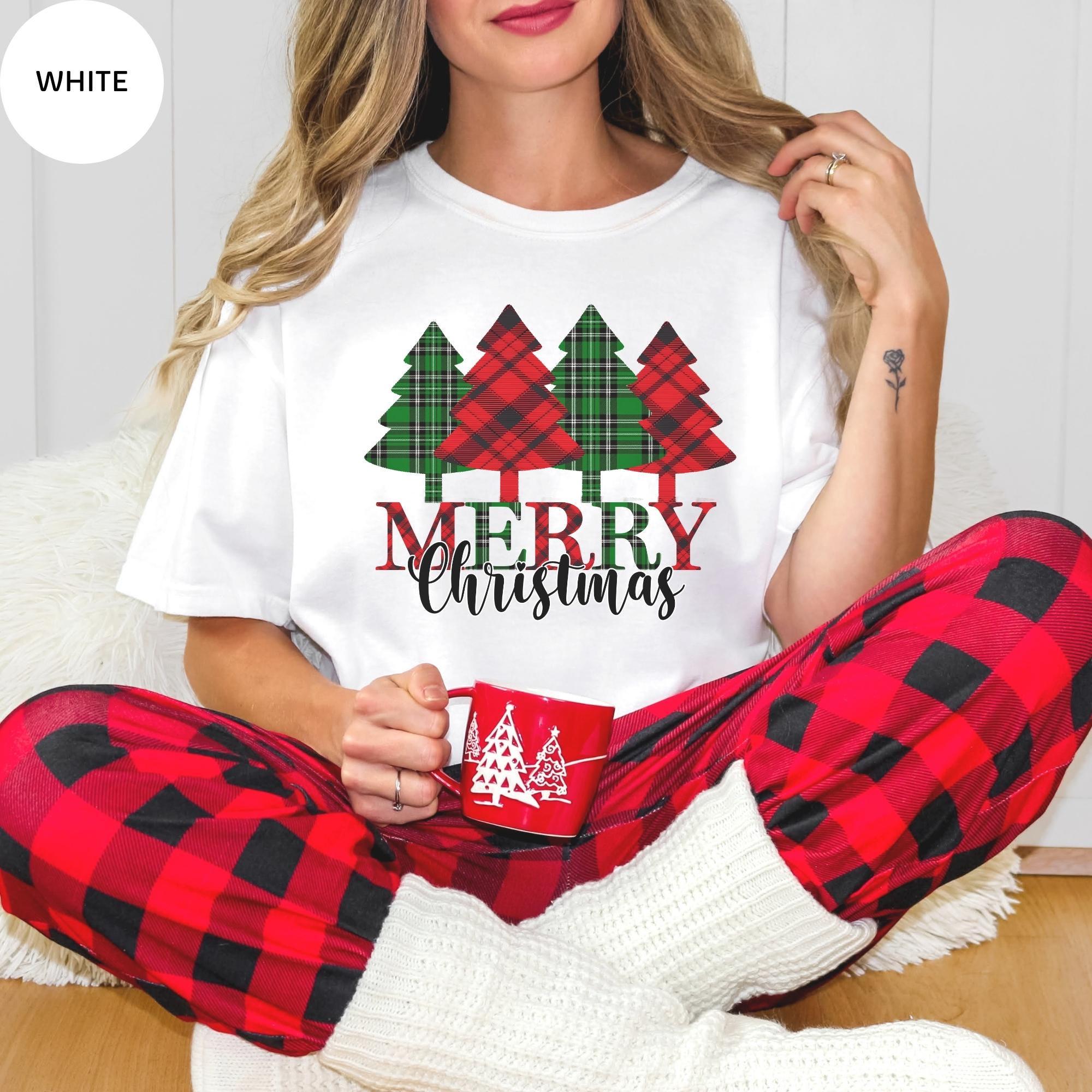 A woman wearing a white t-shirt with a Christmas tree design and the text "Merry Christmas" is sitting on the floor, holding a mug.