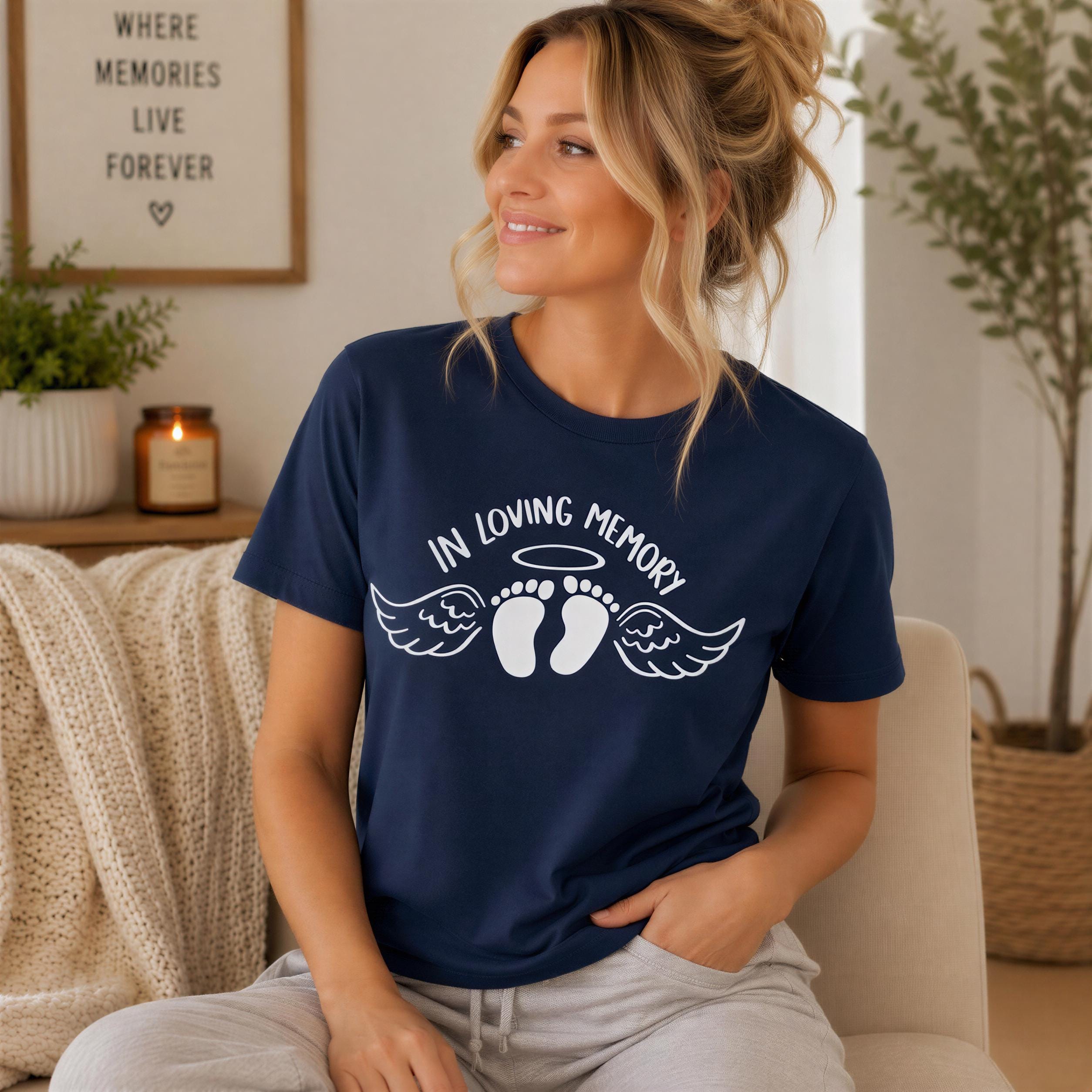 A woman wearing a navy blue t-shirt with a graphic design of wings and baby feet, sitting on a couch in a room with a framed sign that says "WHERE MEMORIES LIVE FOREVER".