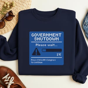 May include: Navy blue sweatshirt with a pixelated graphic that reads "GOVERNMENT SHUTDOWN" with a loading bar at 1%. The text "Please wait..." and "Press Ctrl+Alt-Congress to continue" are also displayed.