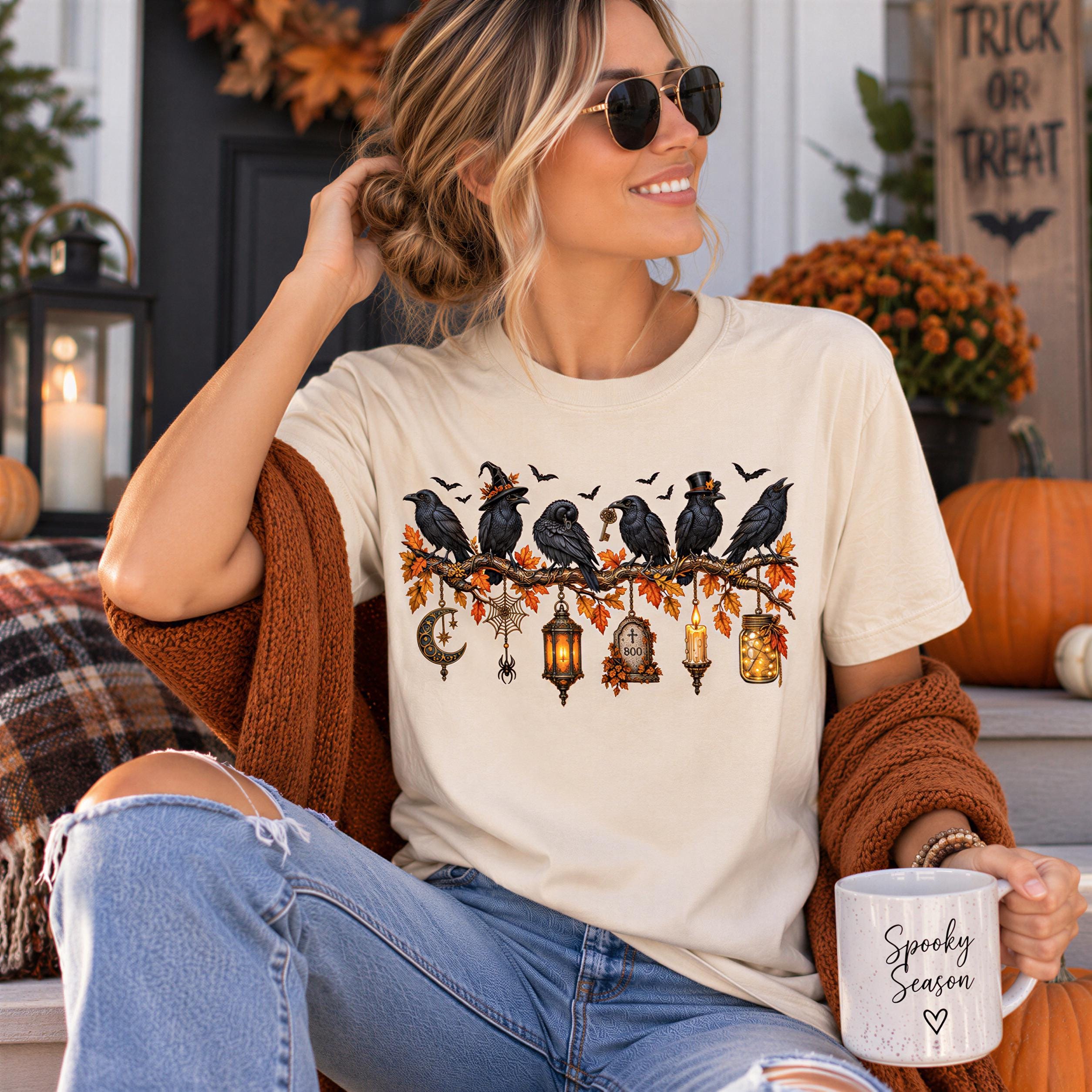 A woman wearing a white t-shirt with a Halloween-themed design, sunglasses, and holding a mug with the text "Spooky Season" is sitting on a porch surrounded by autumn decorations.