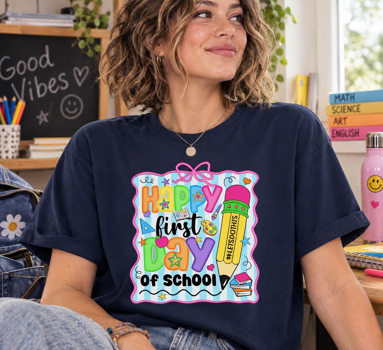 A smiling woman wearing a blue t-shirt with a colorful graphic design that says "Happy first day of school".
