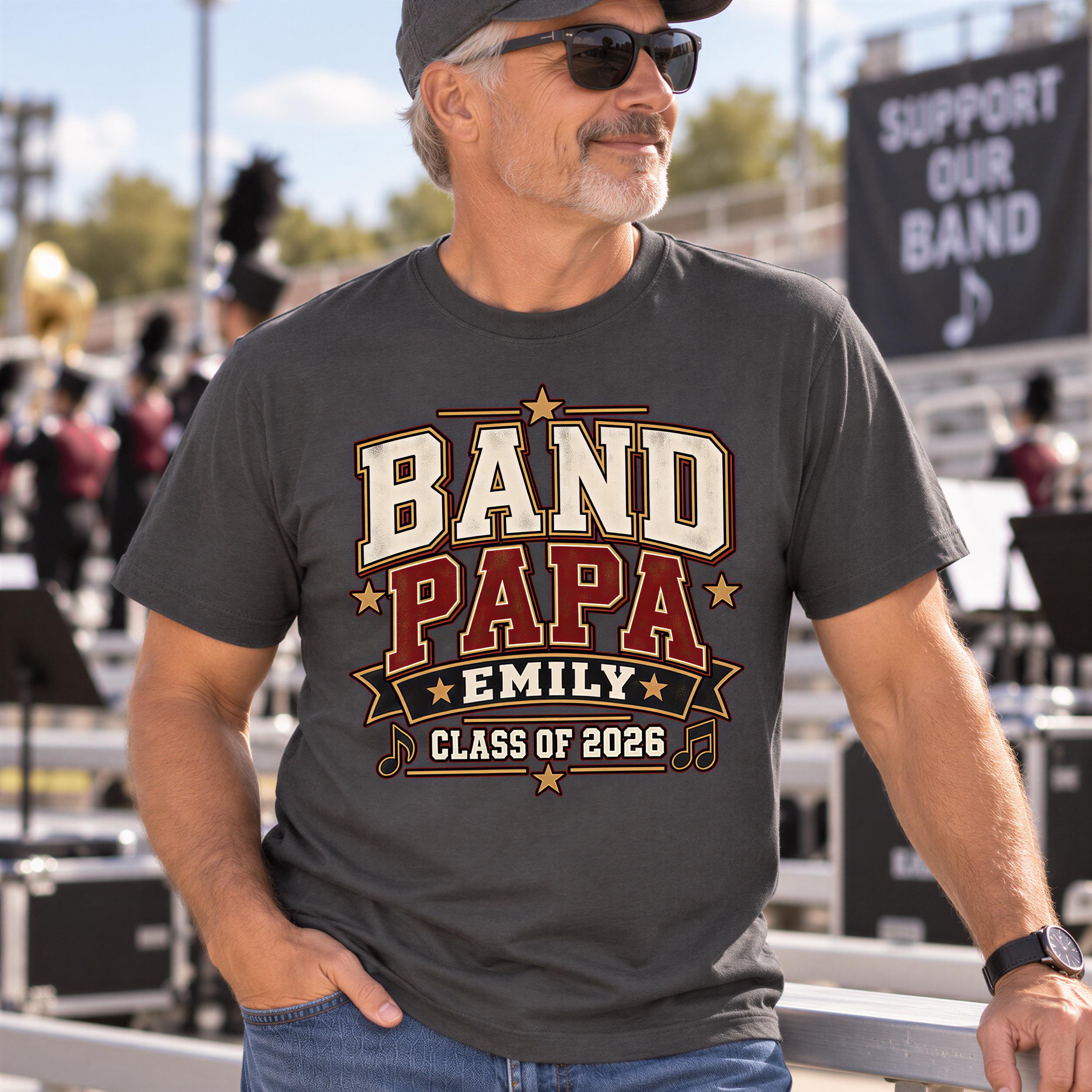 A man wearing a gray t-shirt with a graphic design that says "BAND PAPA EMILY CLASS OF 2026" is standing in front of a crowd.