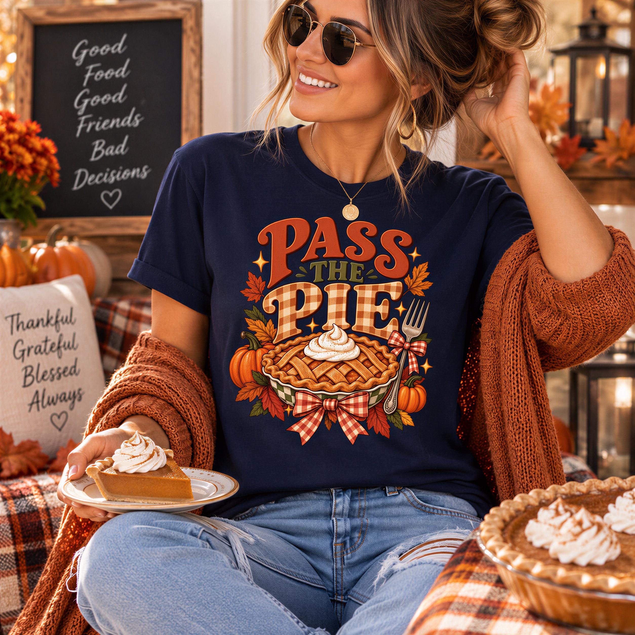 A woman wearing a blue t-shirt with a pumpkin pie graphic and holding a slice of pumpkin pie, smiling and sitting in a cozy autumn setting with a chalkboard sign and other fall decorations.