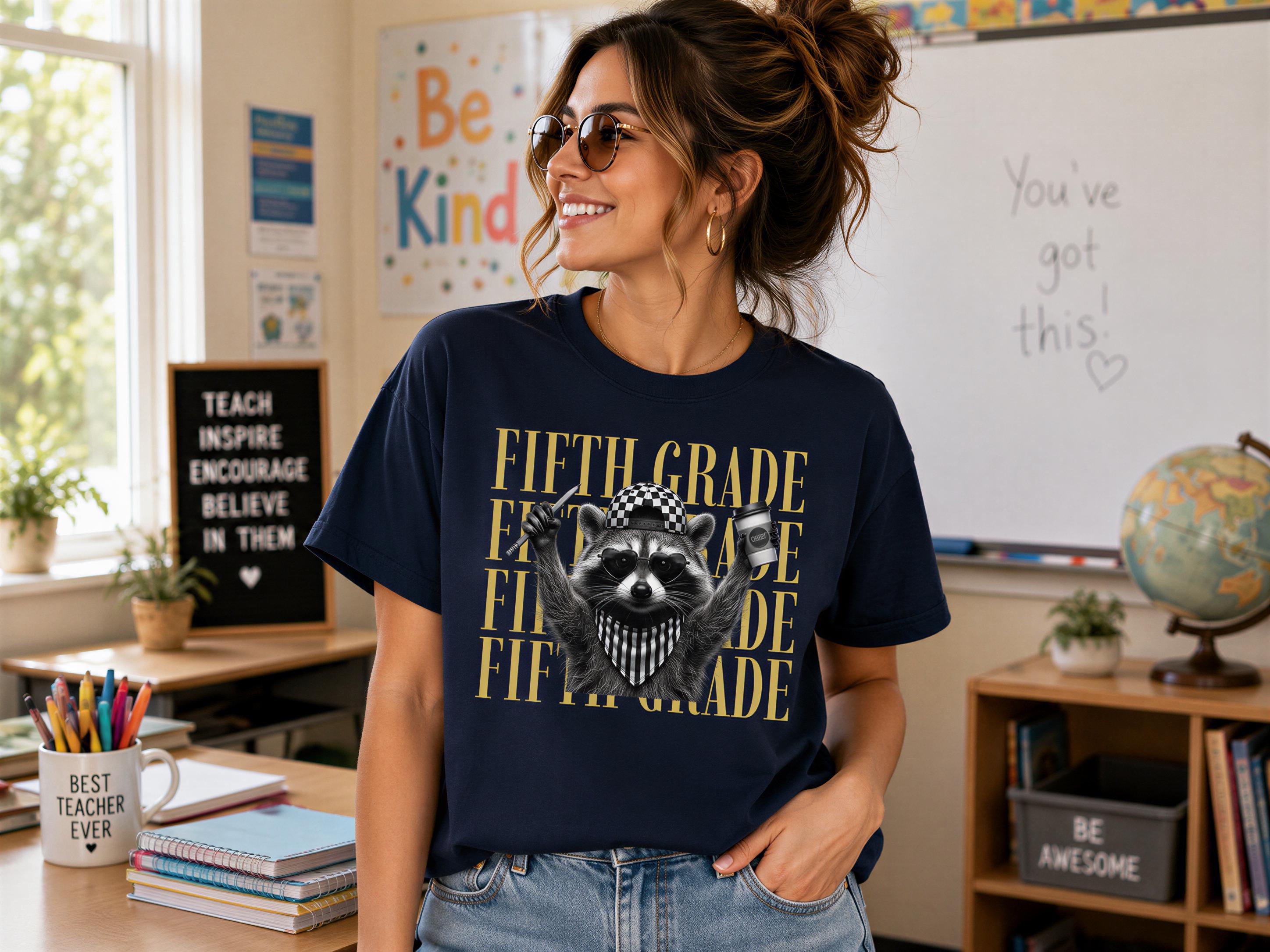 A woman wearing a navy blue t-shirt with a raccoon graphic and text, smiling and standing in a classroom setting with a globe, books, and a bulletin board.