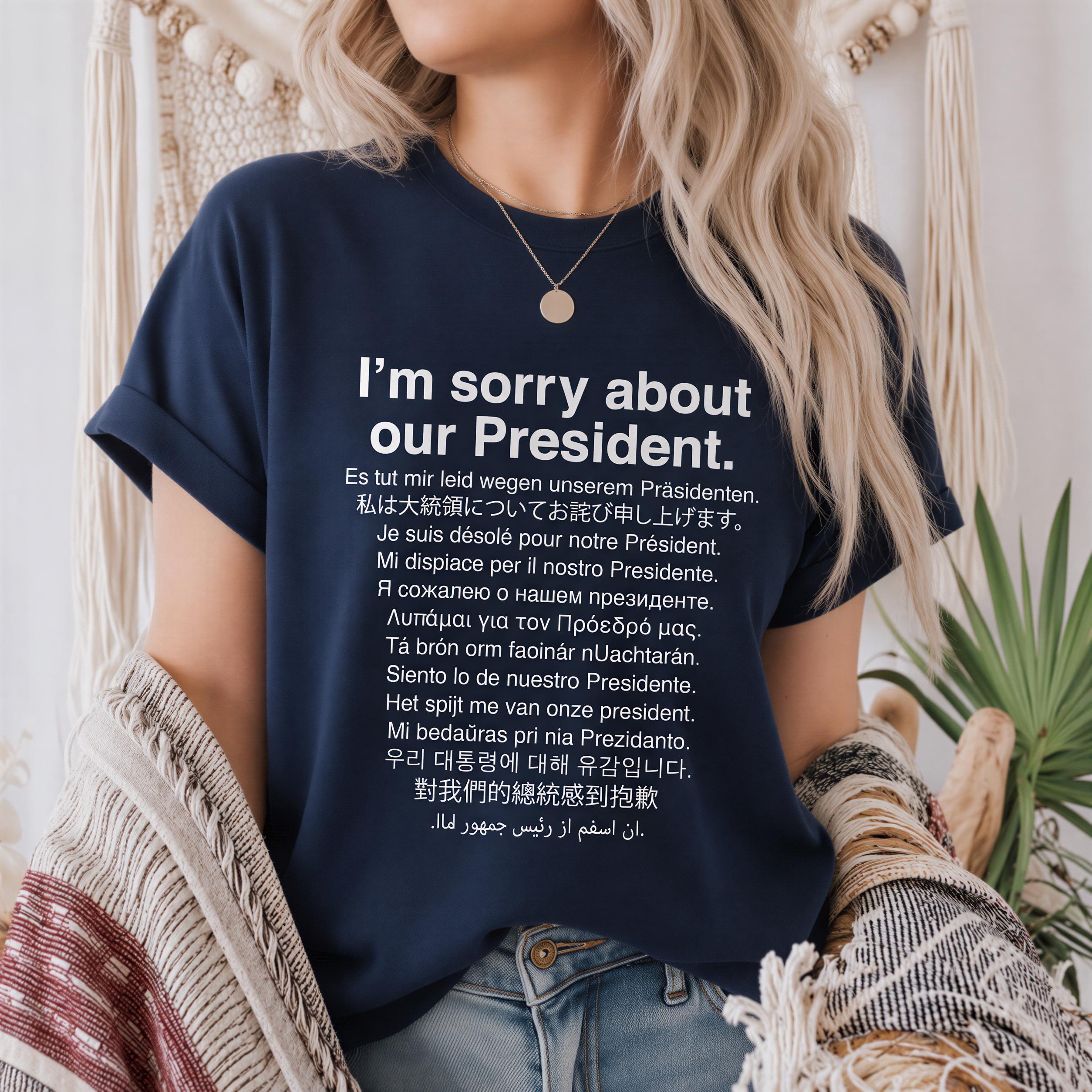 A person wearing a navy blue t-shirt with a message written in multiple languages, including English and Spanish, expressing an apology for the president.