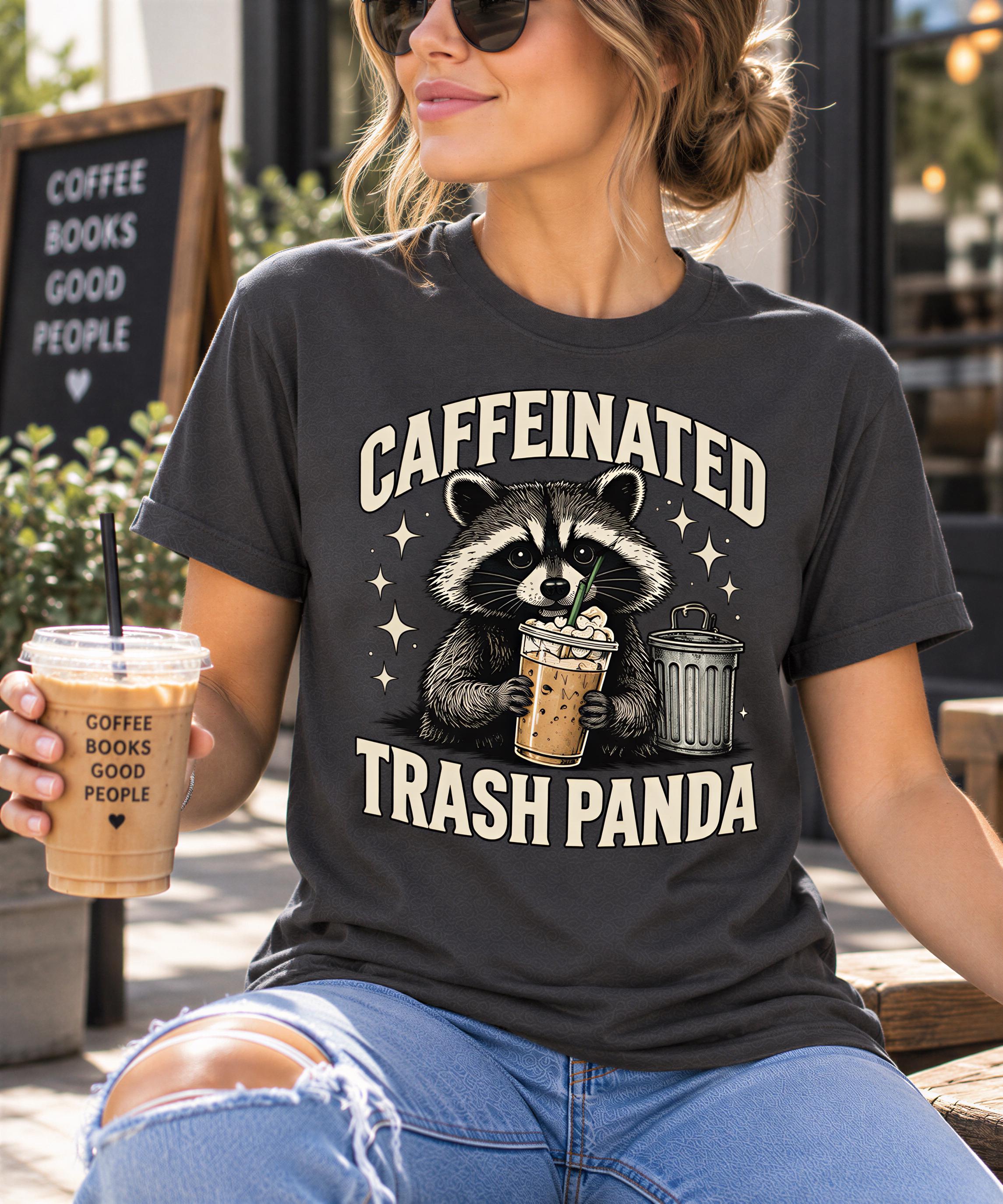 A woman wearing a black t-shirt with a graphic design of a raccoon holding a coffee cup and trash can, sitting outdoors with a drink in her hand.