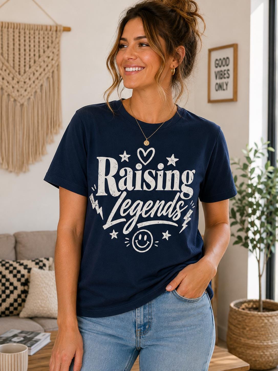 A woman wearing a navy blue t-shirt with the text "Raising Legends" and a smile, standing in a room with a beige wall, a macrame wall hanging, and a potted plant.
