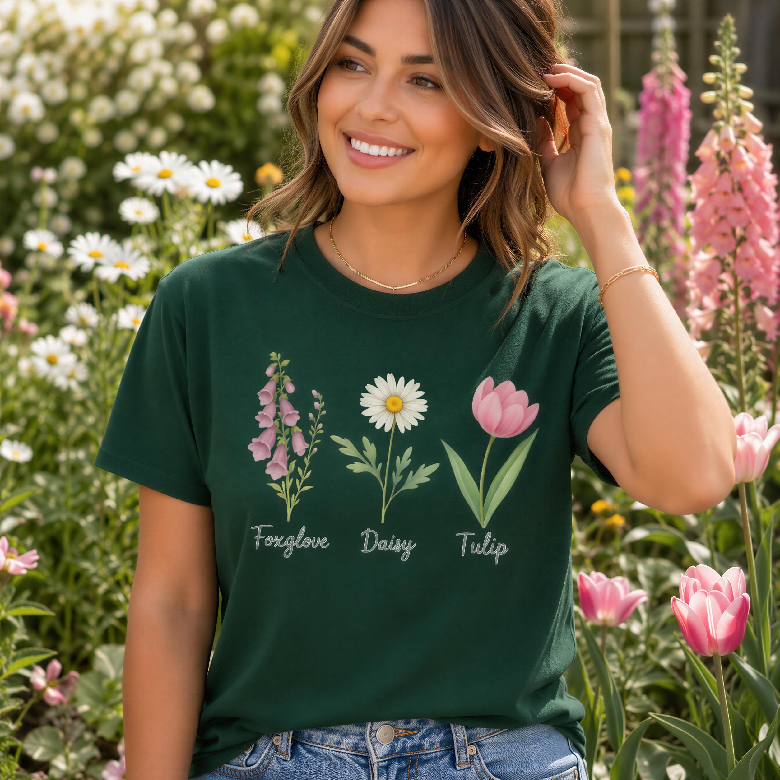 A woman wearing a green t-shirt with illustrations of three flowers: foxglove, daisy, and tulip, standing in a garden surrounded by pink flowers.