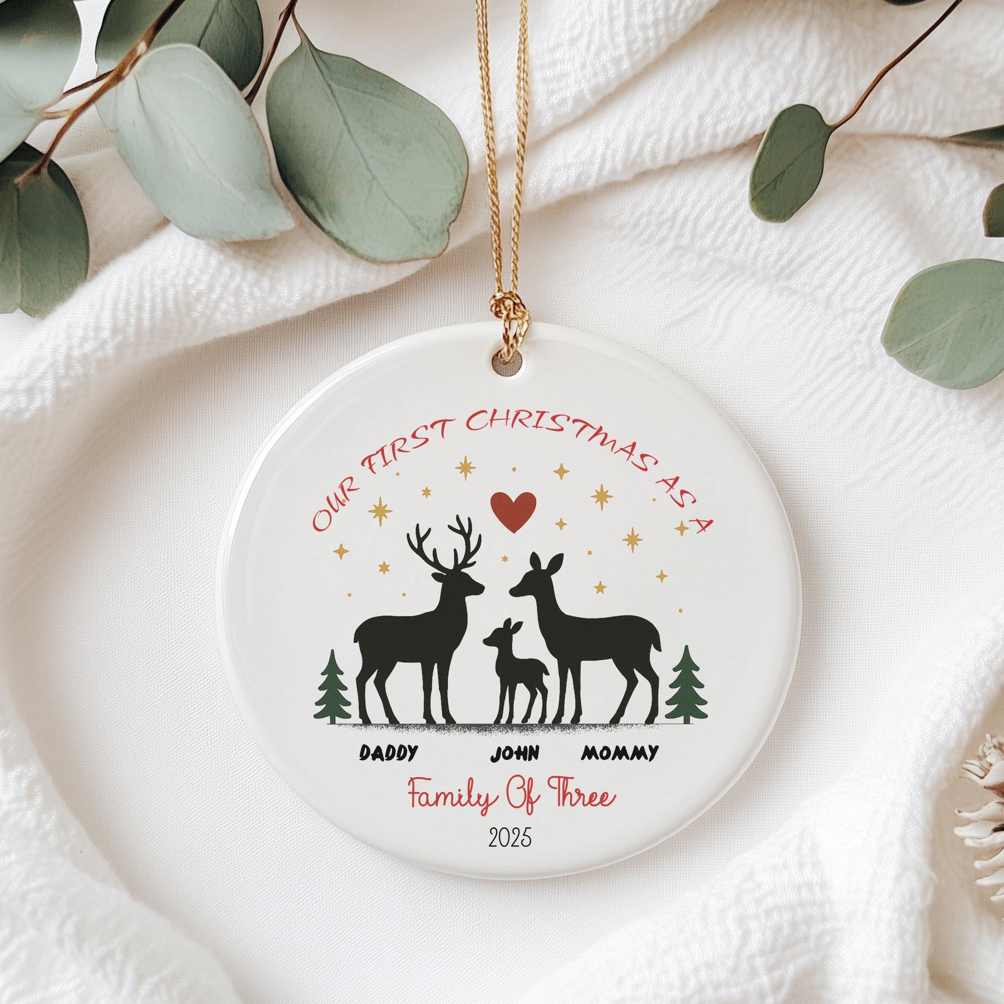 A white ceramic ornament with a design of a family of three deer, including a mother deer, father deer, and their child deer, along with the text "Our first Christmas as a family of three" and the year "2025".