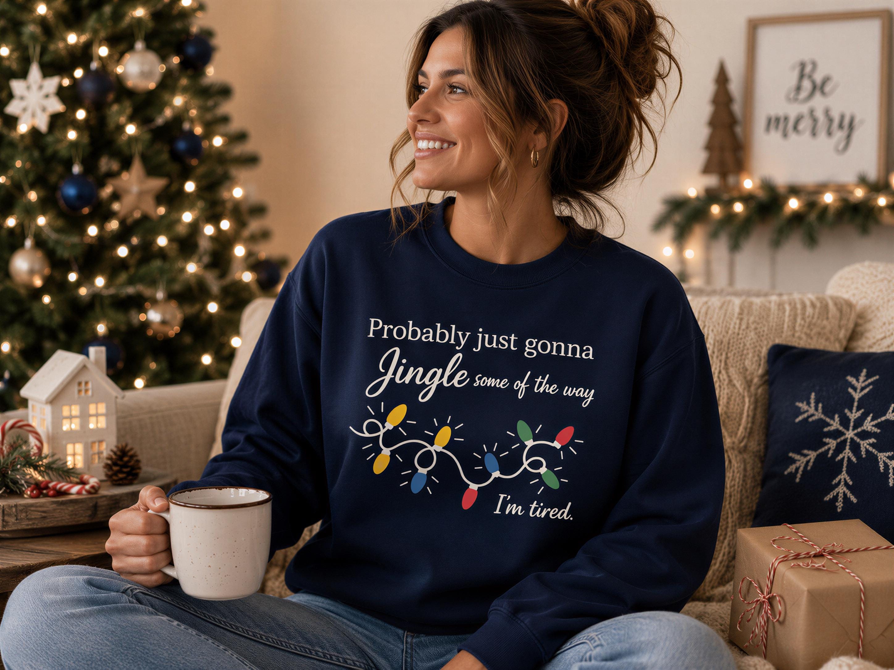 A woman wearing a navy blue sweatshirt with a humorous Christmas-themed design is sitting on a couch, holding a coffee mug and smiling.