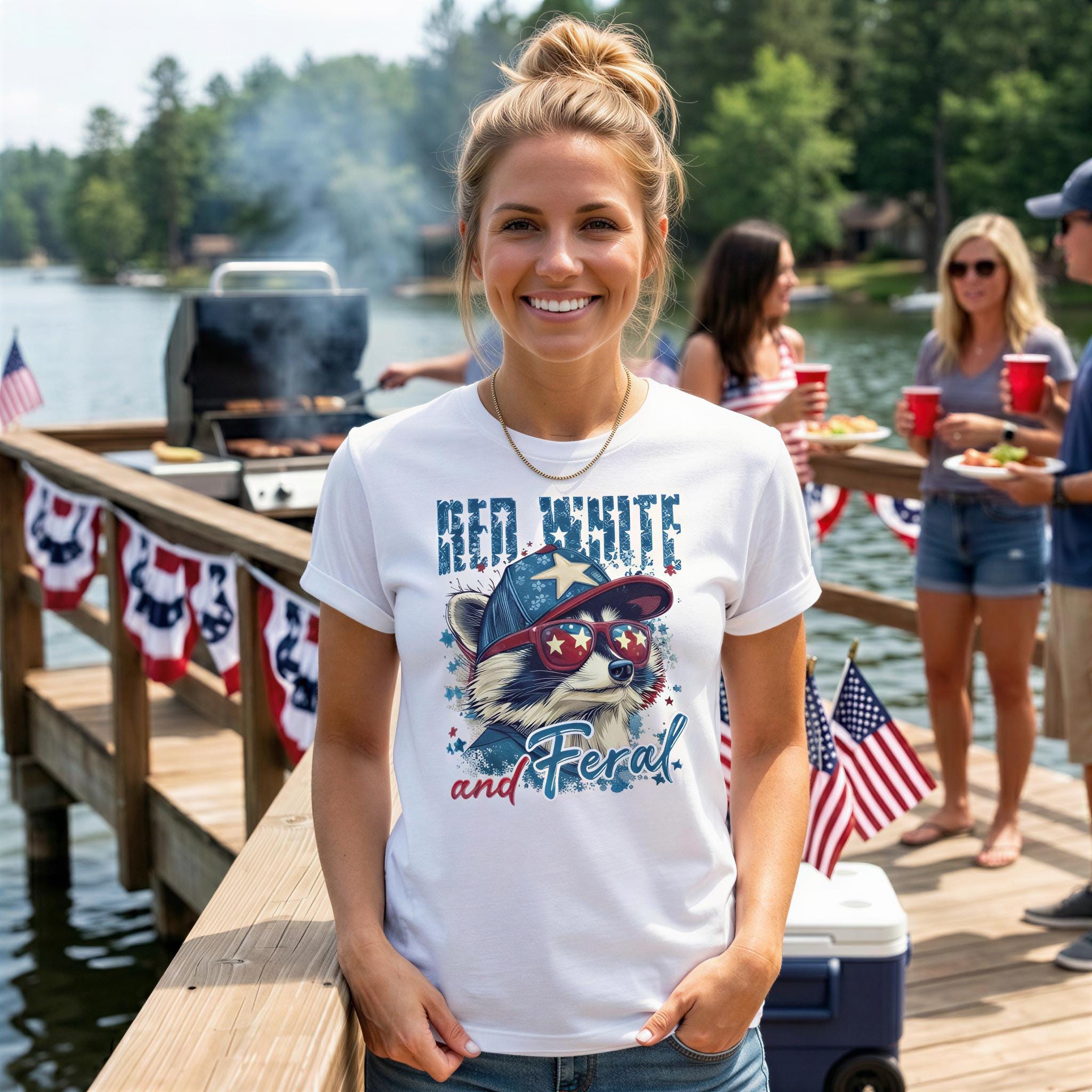 A woman wearing a white t-shirt with a graphic of a raccoon and the text "Red White and Feral" stands on a dock, smiling at the camera. In the background, there are other people and a grill, suggesting a social gathering or outdoor event.
