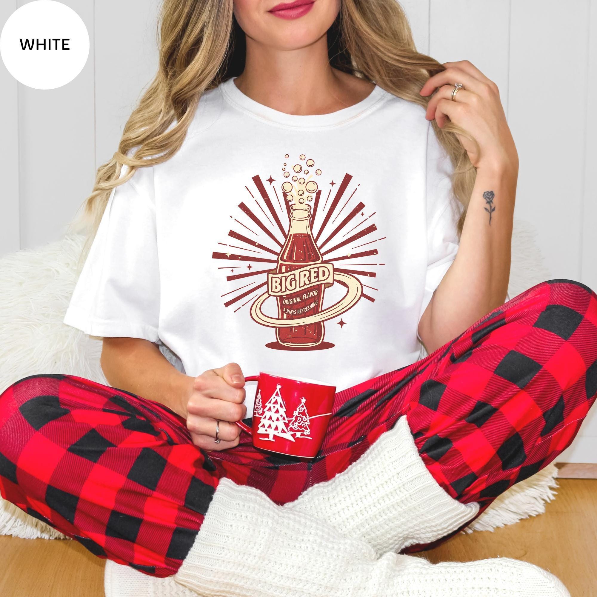 A woman wearing a white t-shirt with a graphic design and red plaid pajama bottoms, holding a red mug, and sitting on the floor.