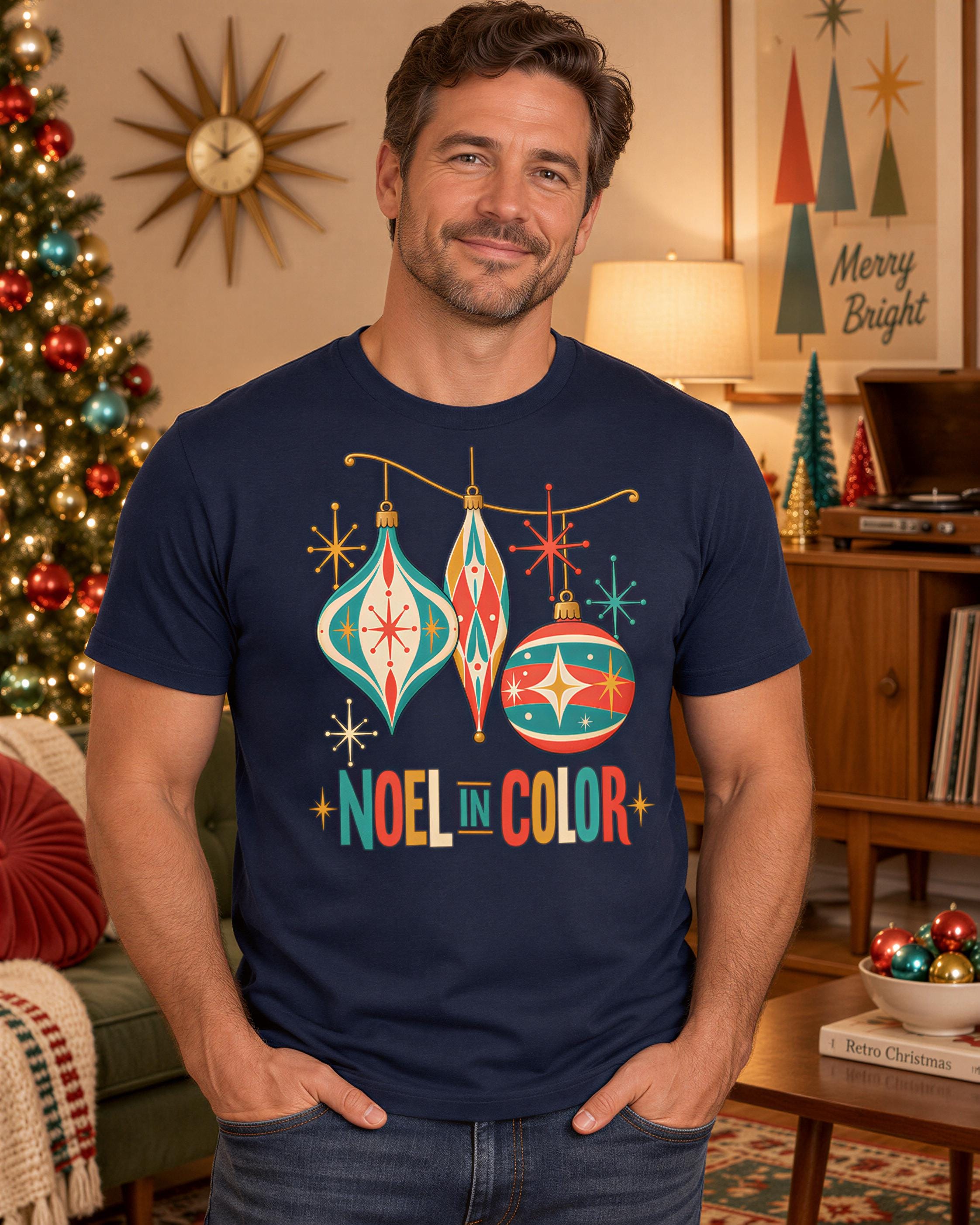 A man wearing a navy blue t-shirt with a colorful graphic design of Christmas ornaments and the text "Noel in Color" stands in front of a Christmas tree and other holiday decorations.