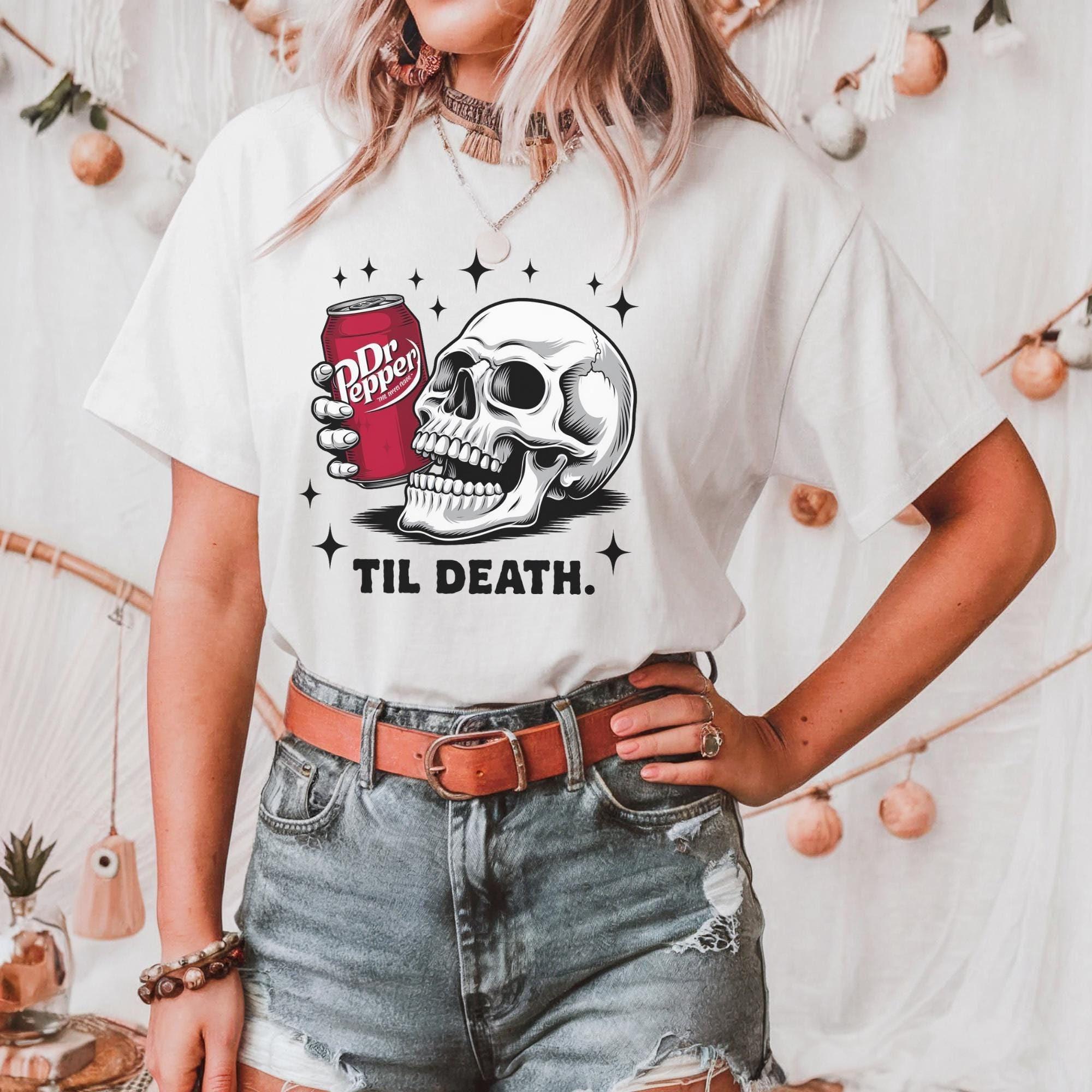 A person wearing a white t-shirt with a graphic design of a skull holding a can of Dr Pepper, accompanied by the text "Til Death."