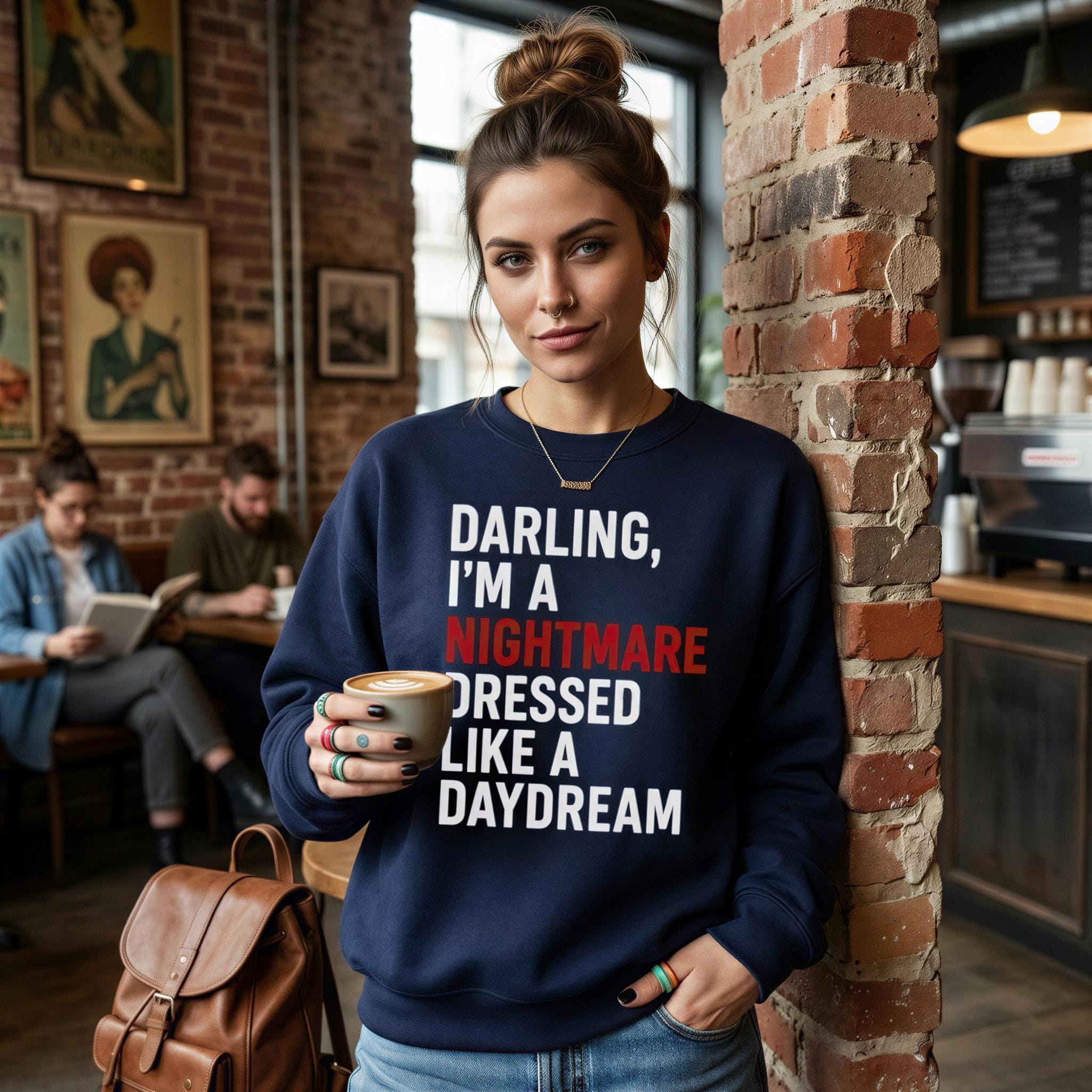 A woman wearing a navy blue sweatshirt with the text "DARLING, I'M A NIGHTMARE DRESSED LIKE A DAYDREAM" stands in a cafe, holding a cup of coffee.