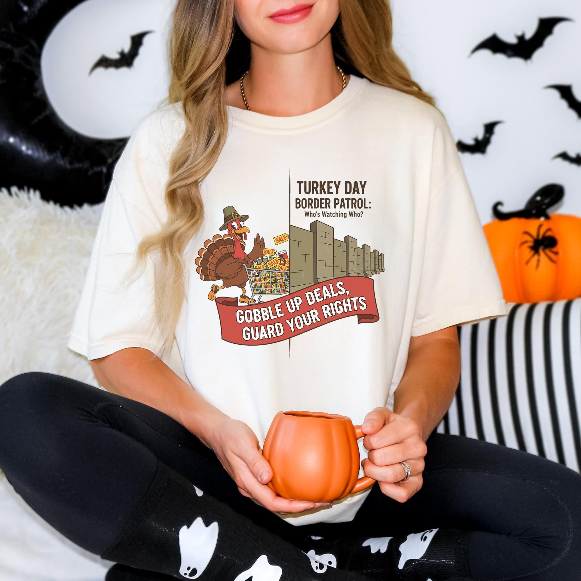 A woman wearing a white t-shirt with a graphic design of a turkey holding a sign that says "Gobble up your rights" and "Border patrol: Who's watching who". She is sitting on a couch with Halloween decorations in the background.