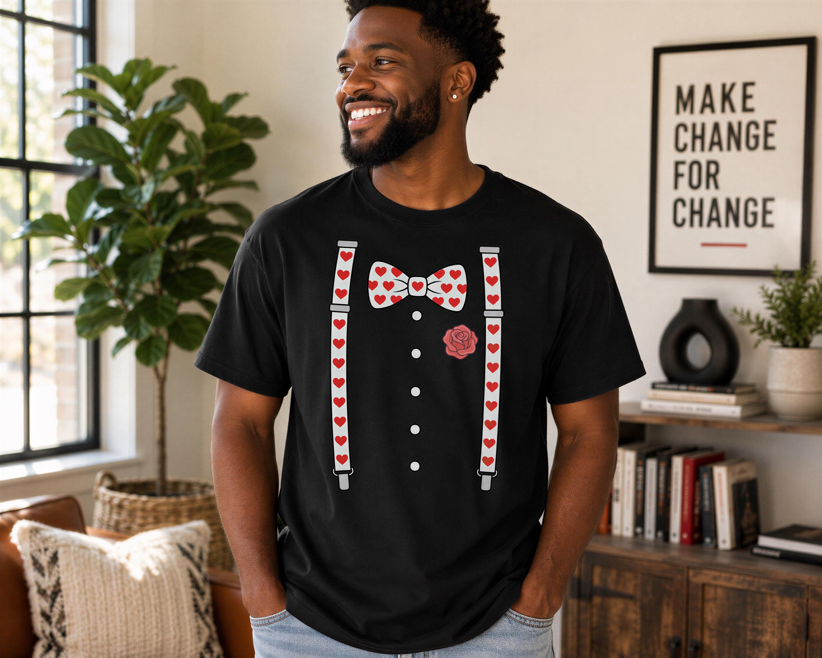 A man wearing a black t-shirt with a bow tie and suspenders printed on it, smiling and standing in a room with a window and plants.
