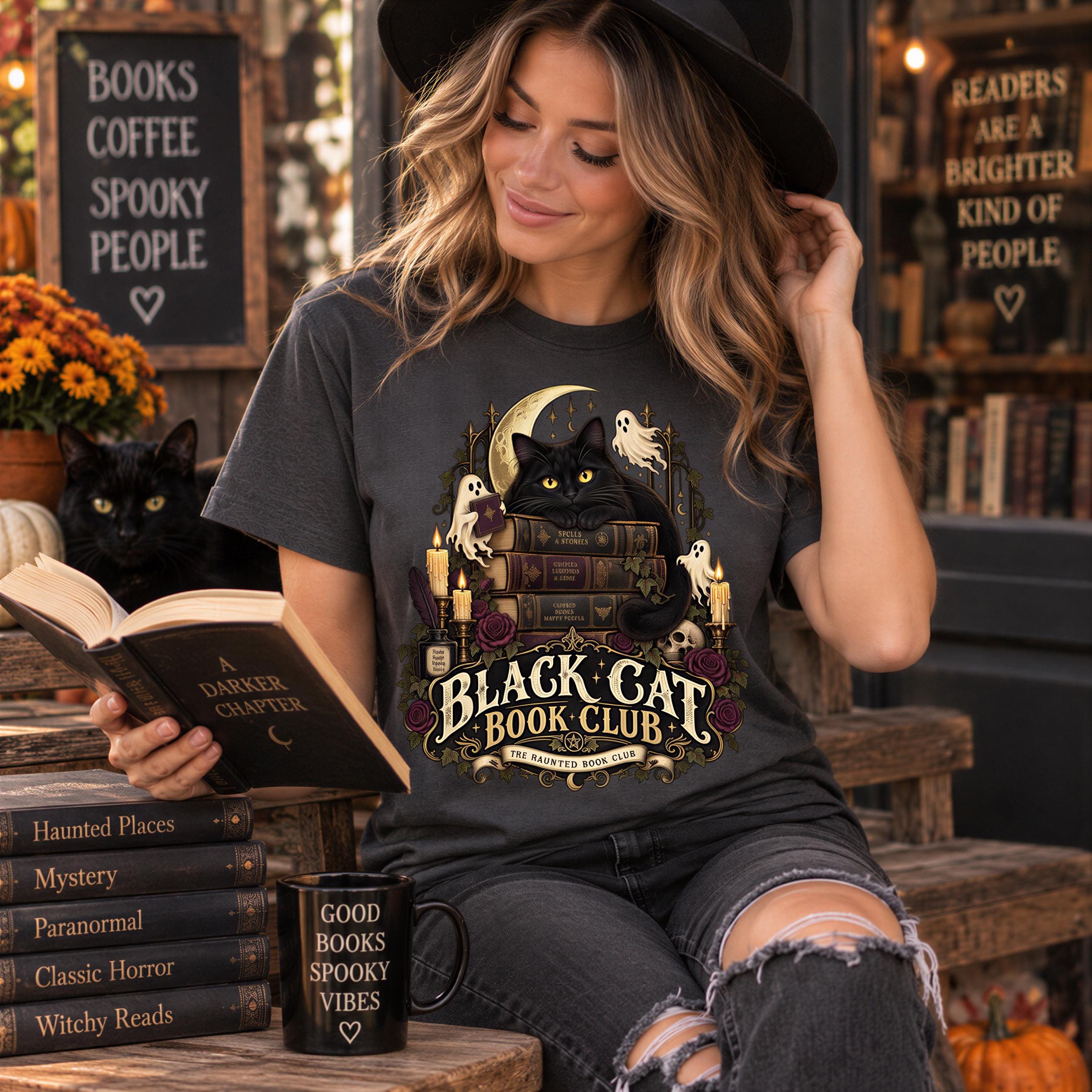 A woman wearing a black hat and a t-shirt with a black cat graphic is sitting on a bench and reading a book. There are several books and a mug with a black cat graphic on a table in front of her.