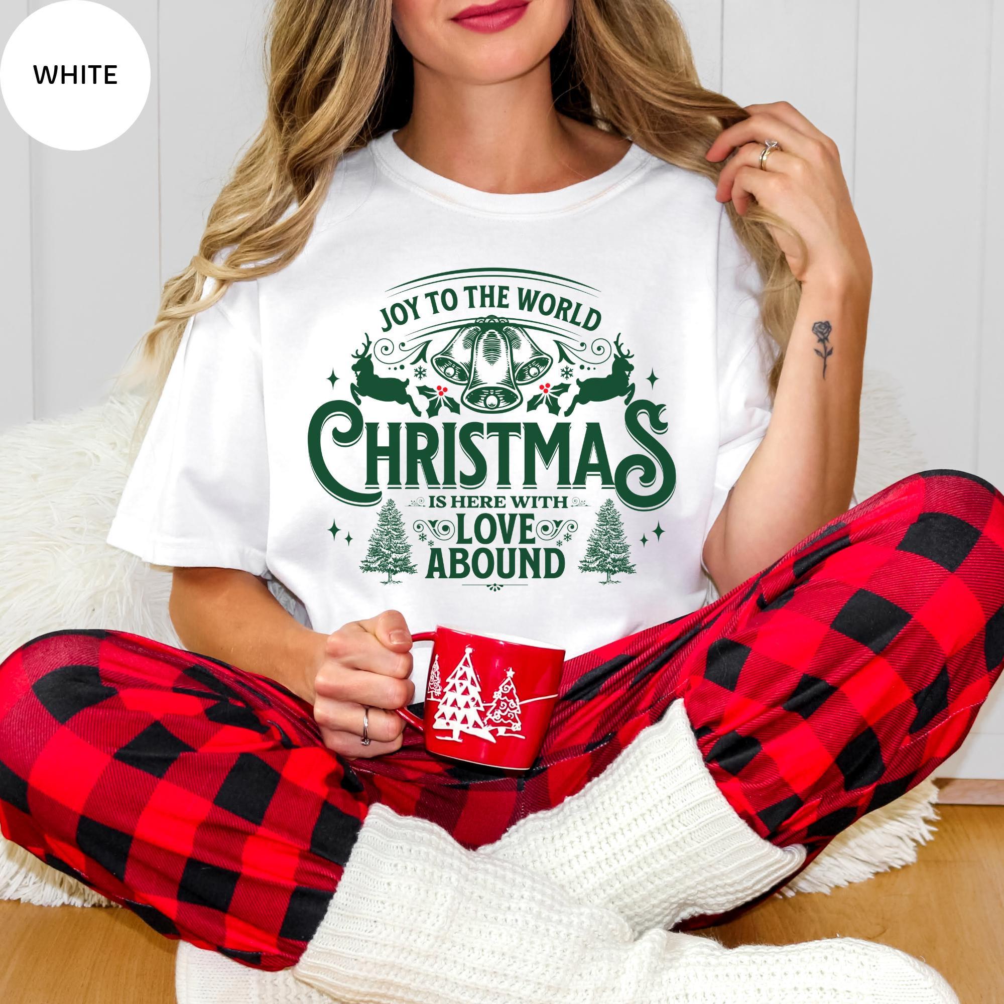 A woman wearing a white t-shirt with a Christmas-themed design and holding a red mug, sitting on the floor with her legs crossed.