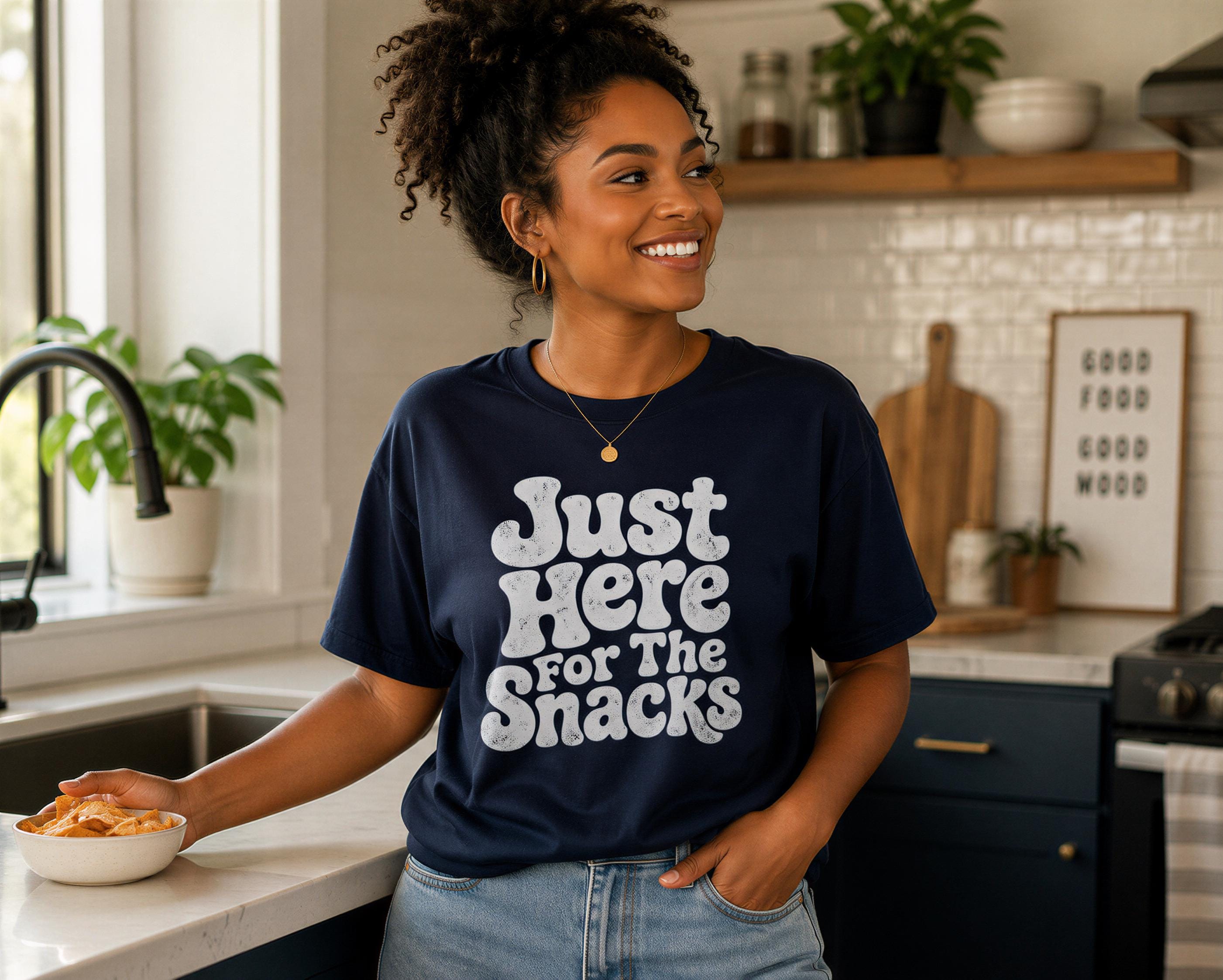A woman wearing a navy blue t-shirt with the text "Just Here for the Snacks" is smiling and holding a bowl of food in a kitchen setting.