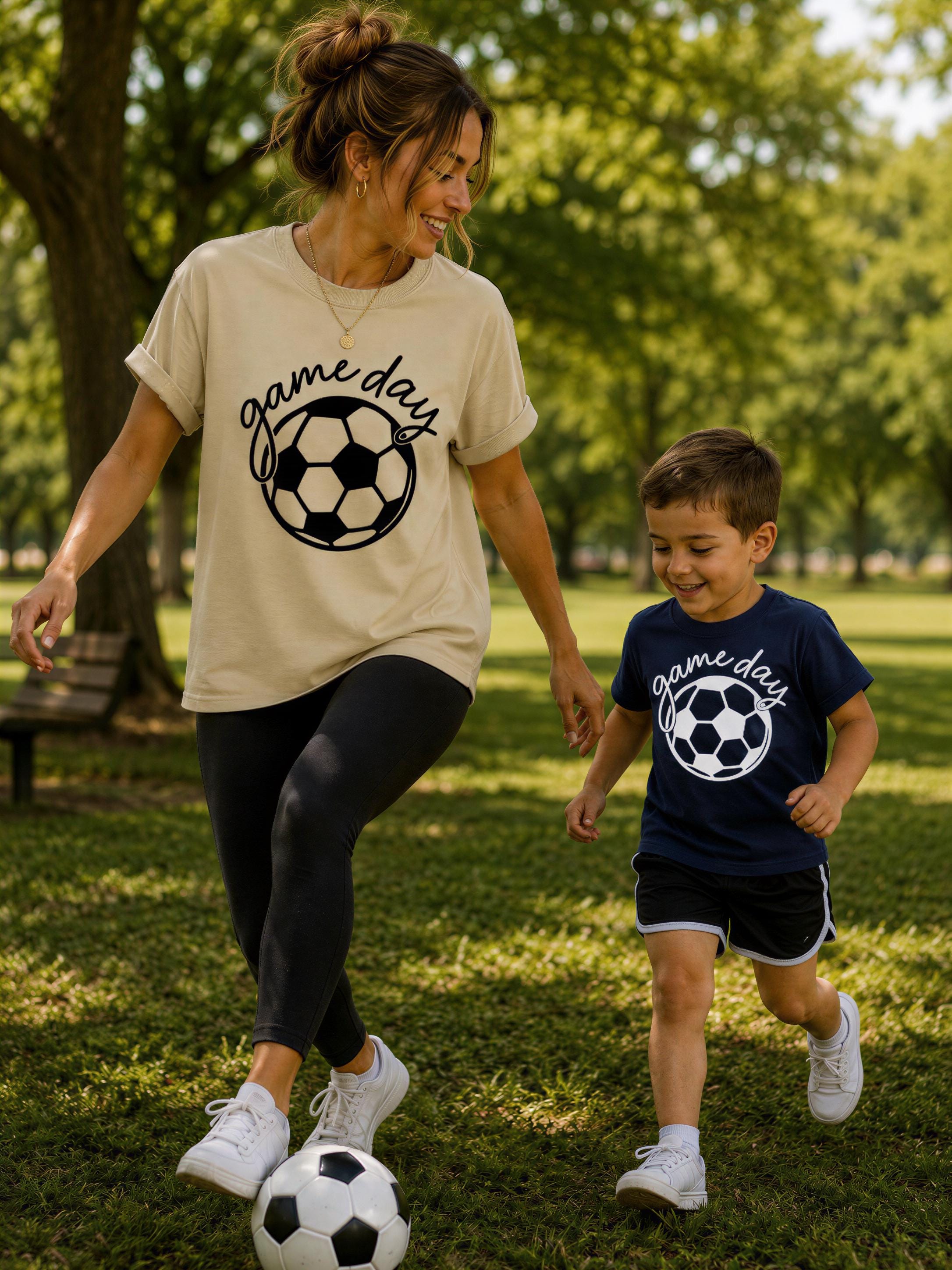 A woman and a young boy are playing soccer in a park, both wearing matching t-shirts with the text "game days" printed on them.