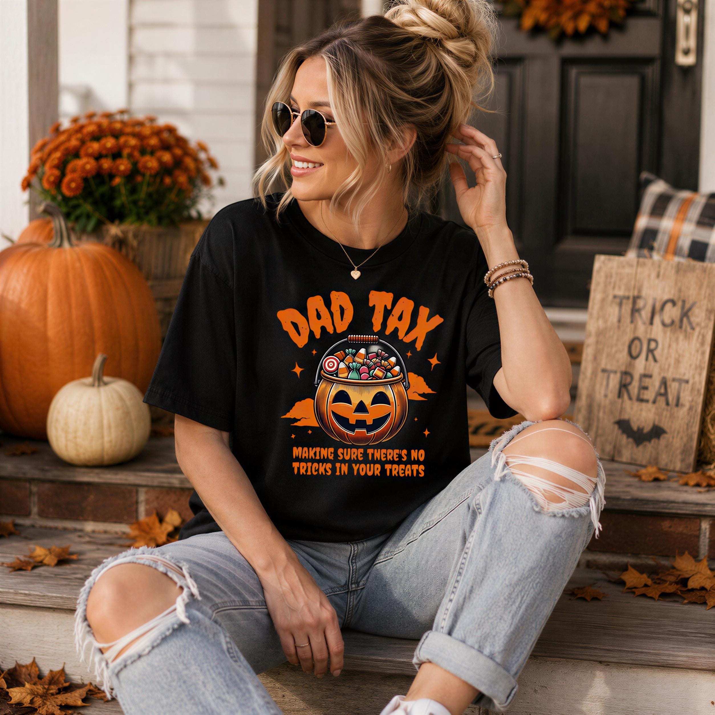 A woman wearing a black t-shirt with a pumpkin graphic and the text "DAD TAX" is sitting on a porch surrounded by autumn decorations, including pumpkins and a "TRICK OR TREAT" sign.